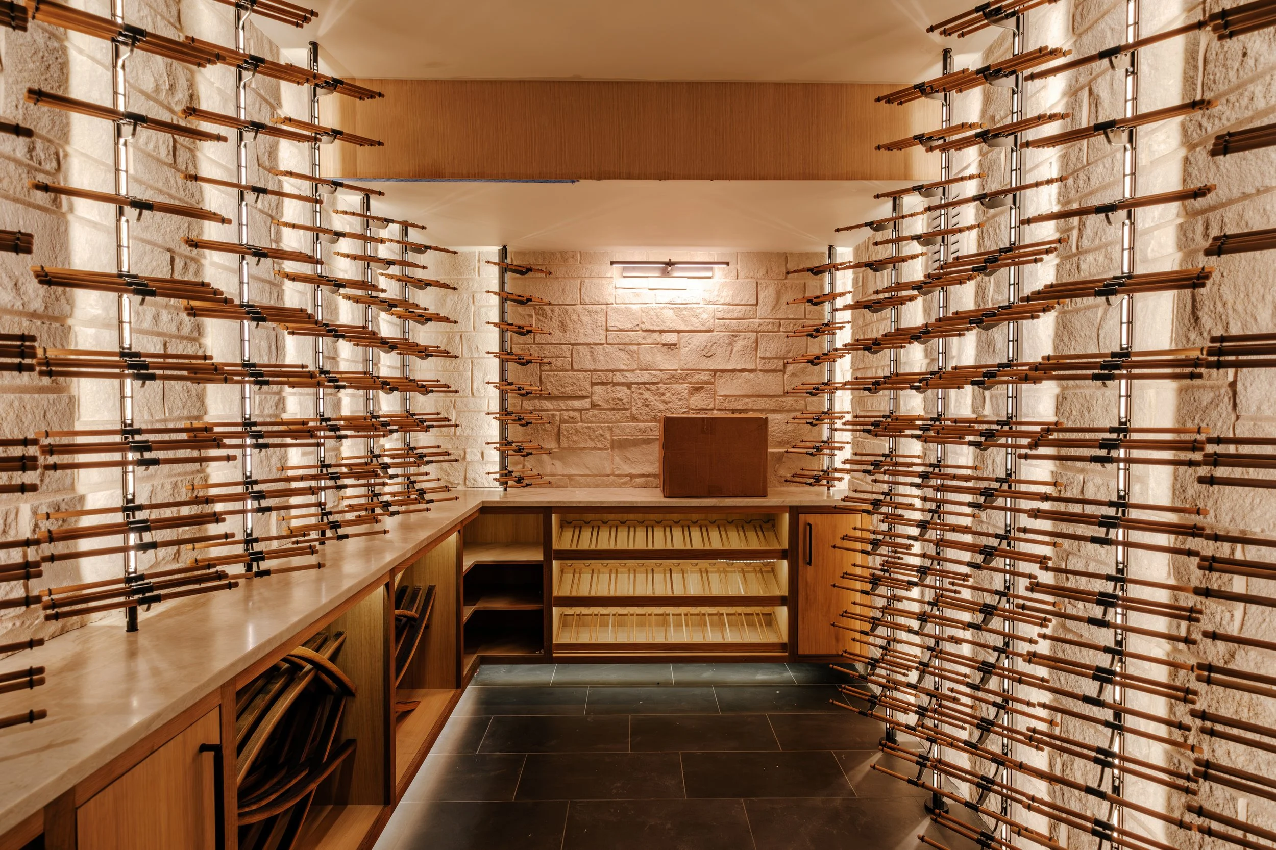 A room with walls made of stone bricks, featuring wooden shelves and multiple rows of copper pipes with black fittings arranged on the walls and ceiling, and a wooden countertop with open compartments underneath.