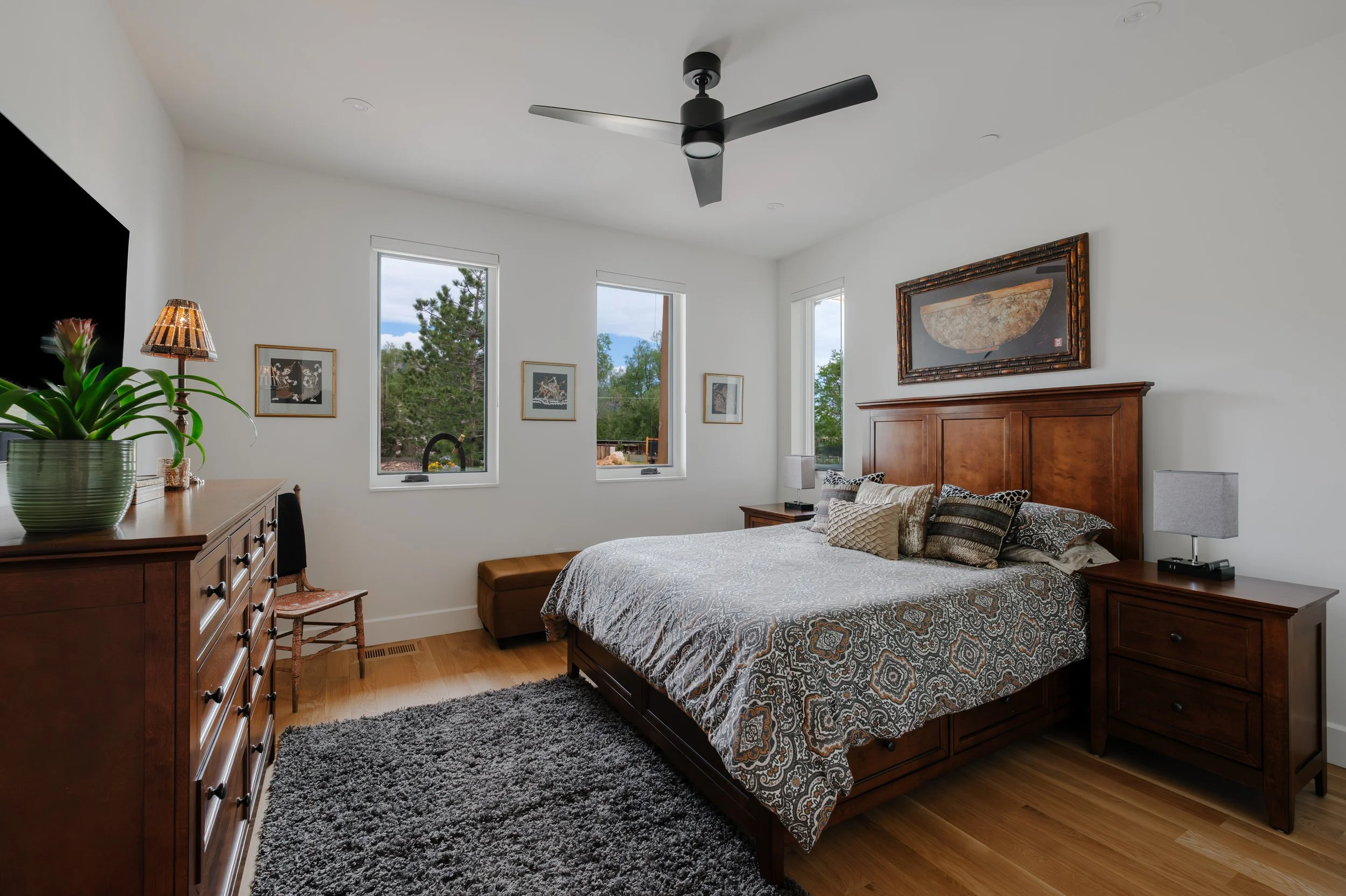 A bedroom with a wooden bed frame, patterned bedding, multiple framed pictures on white walls, two windows showing trees outside, a wooden dresser with a green plant, lamps, and a ceiling fan.