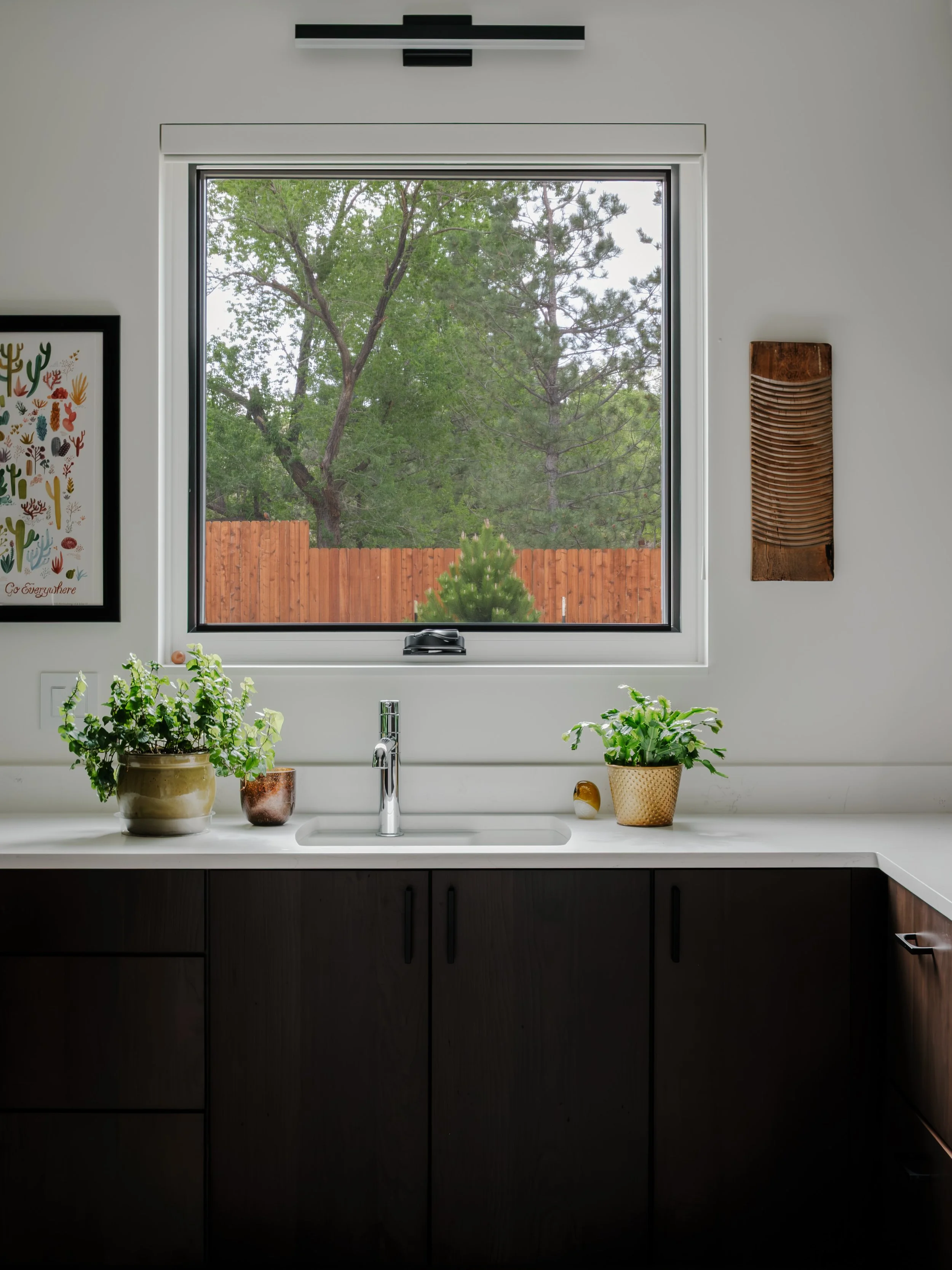 A view of a kitchen window with a white frame, overlooking a backyard with green trees and a wooden fence. The white kitchen sink with a silver faucet is below the window, with potted plants on the countertop and decorative items on the wall.