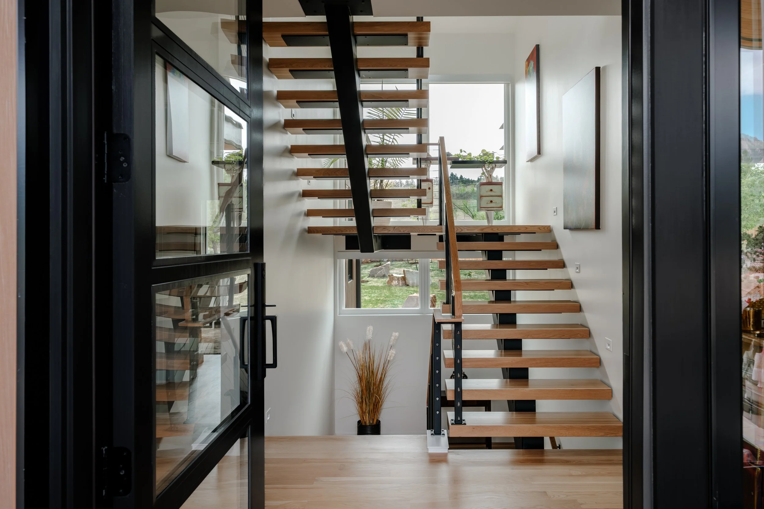 View of an interior stairway with wooden steps and black metal railing, situated next to large windows overlooking a green outdoor landscape.