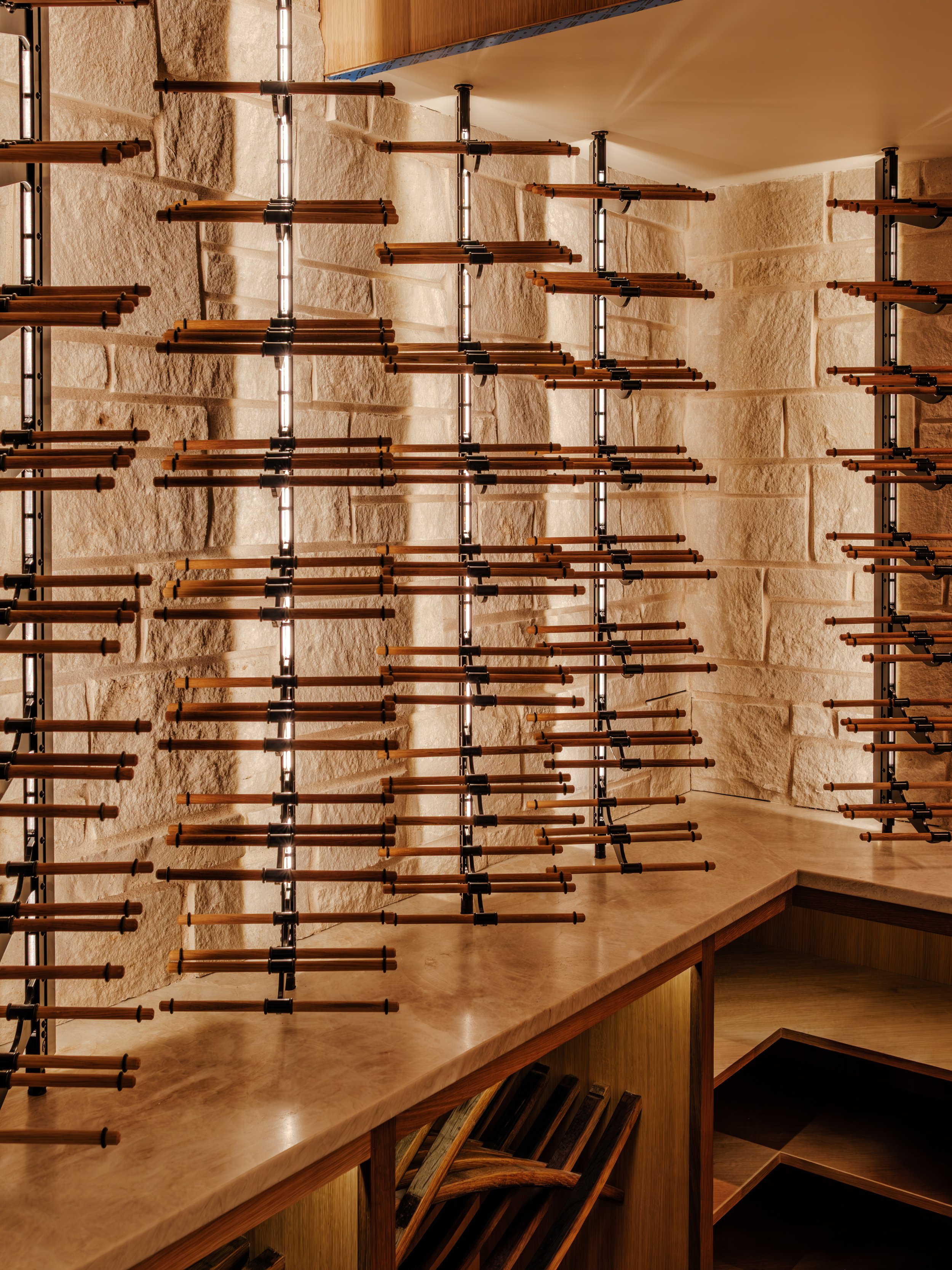 Interior of a wine cellar with wooden wine racks against a textured stone wall, illuminated by vertical light strips and a wooden countertop below the racks.