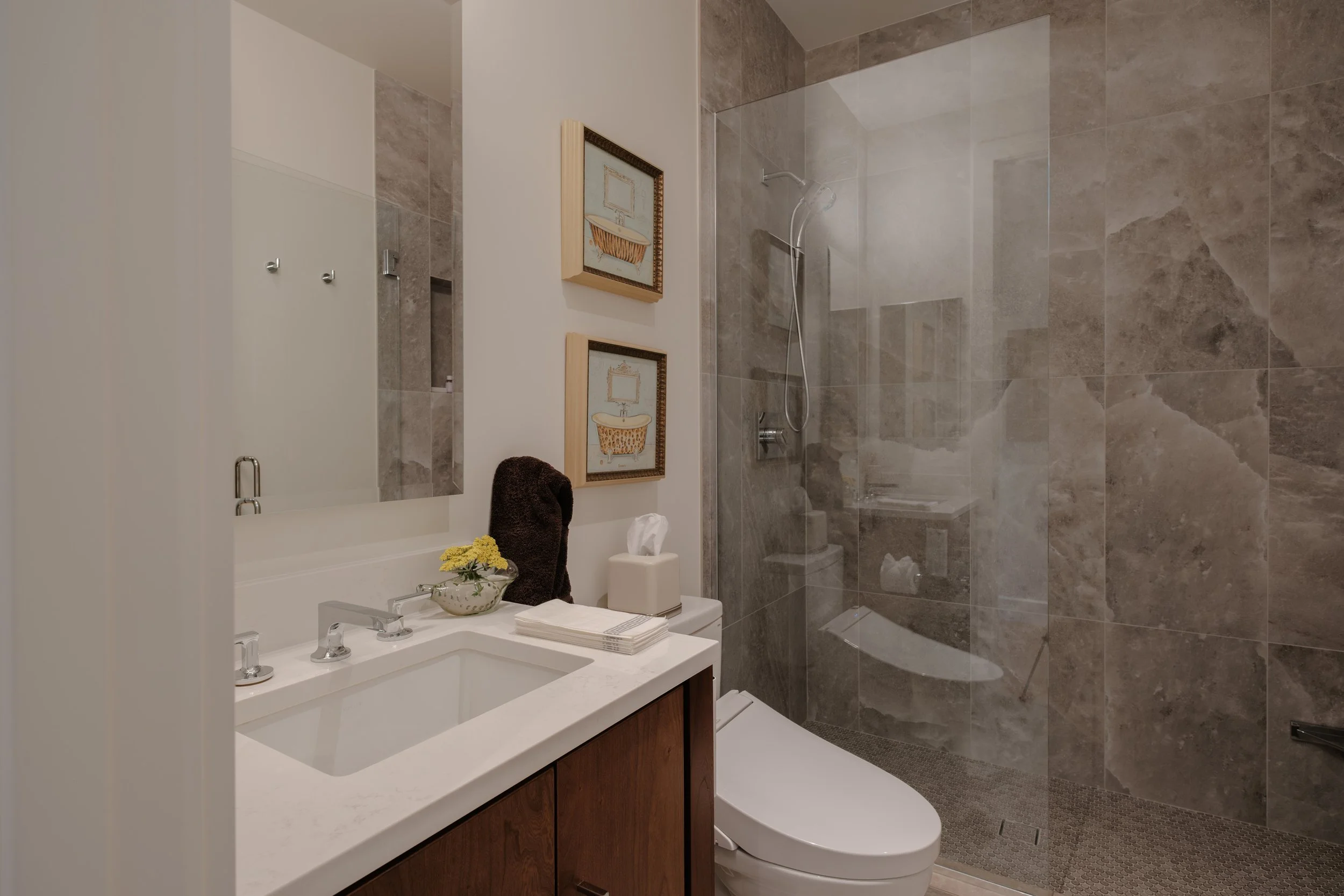 A modern bathroom with a vanity mirror, sink, and faucet, decorated with framed artwork and a small flower arrangement. A toilet is visible in front of the vanity. The shower area features large beige marble tiles, a rain showerhead, and a glass part