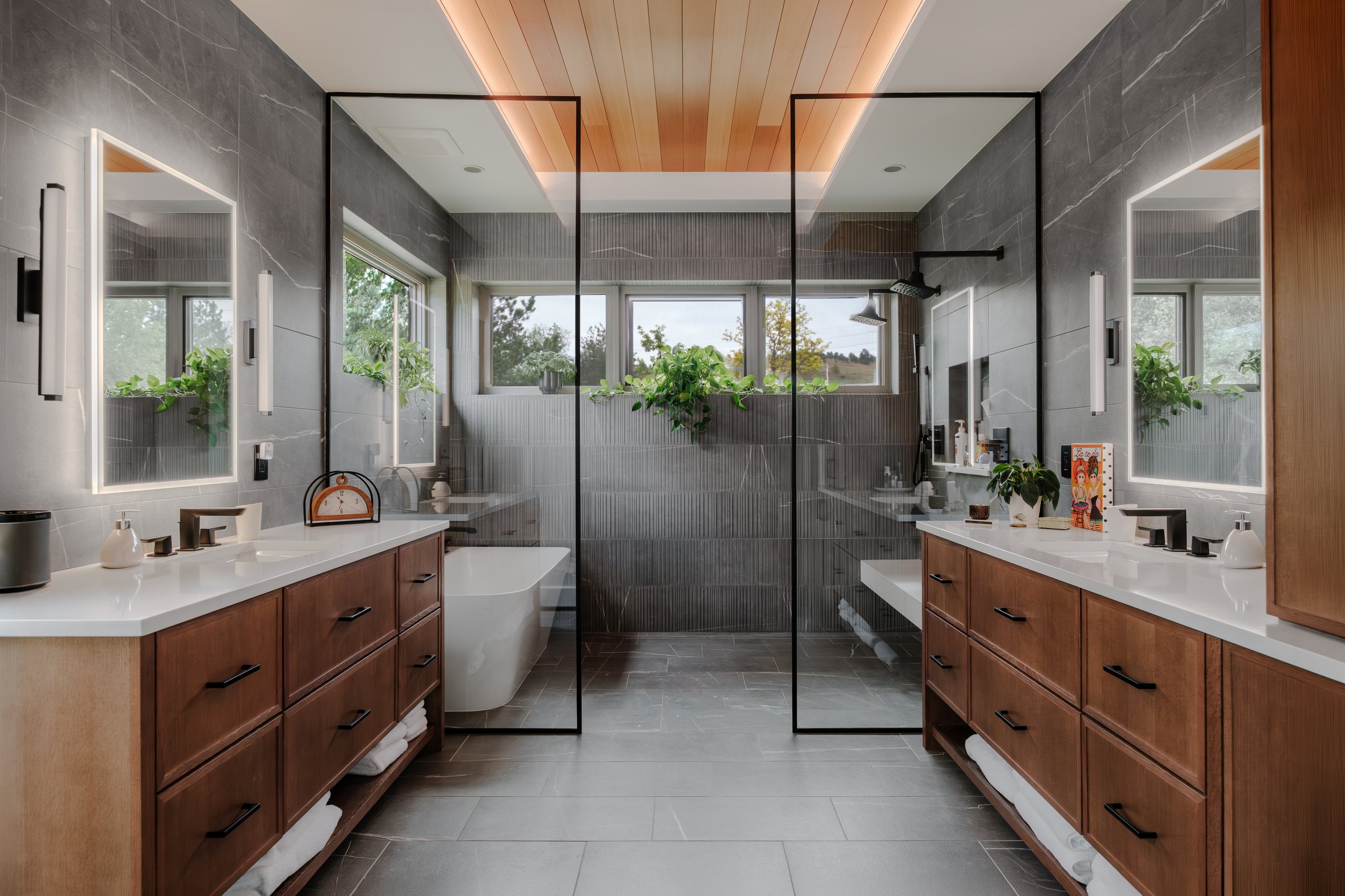 Modern bathroom with double vanity sinks, large mirrors with LED lights, gray marble tiles, and a glass shower enclosure with a black frame. Green plants are on the windowsills, and there is a view of trees outside.