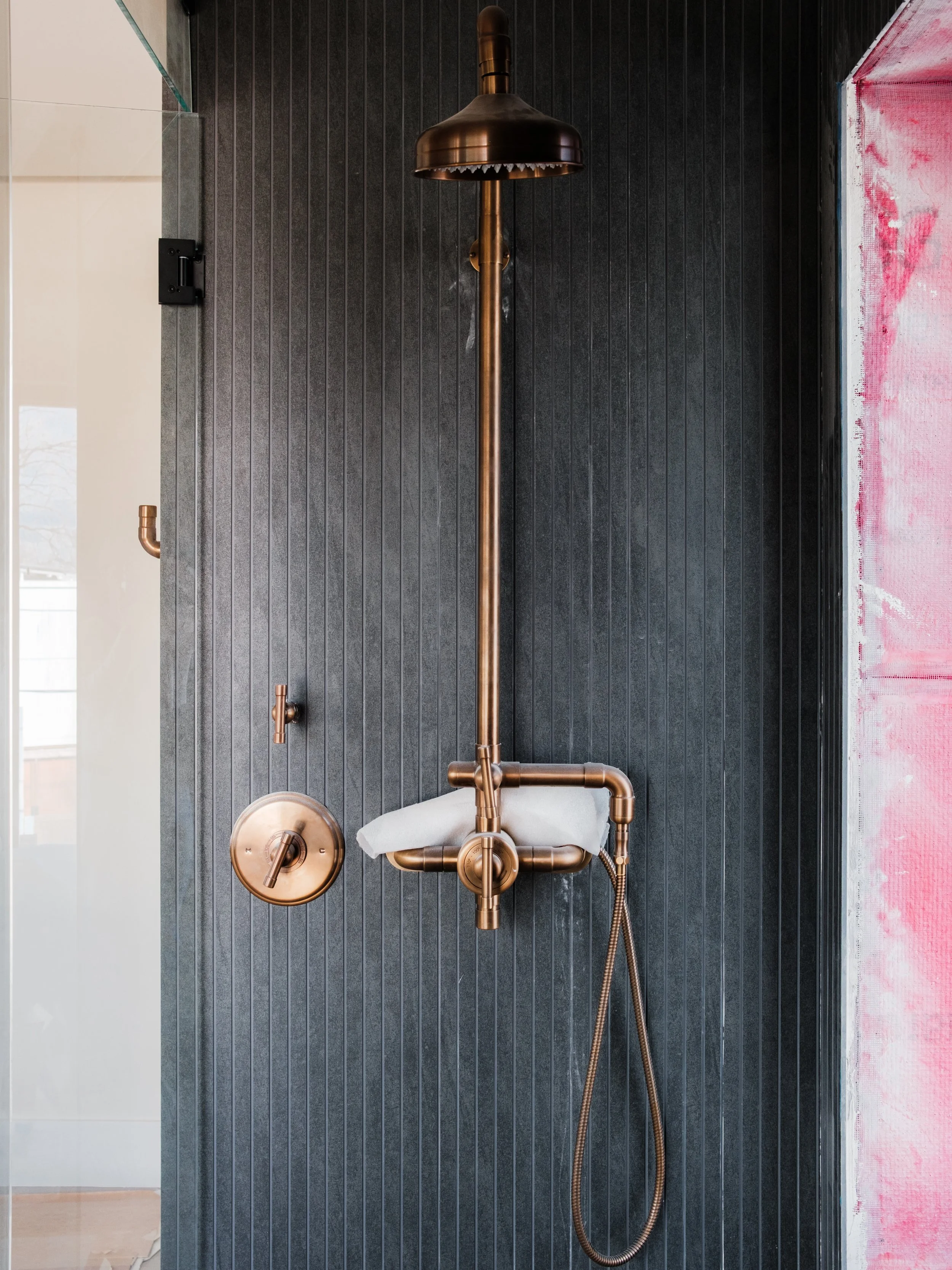 Bronze shower fixture with a large round showerhead, control knobs, and a handheld showerhead mounted on a black textured wall.