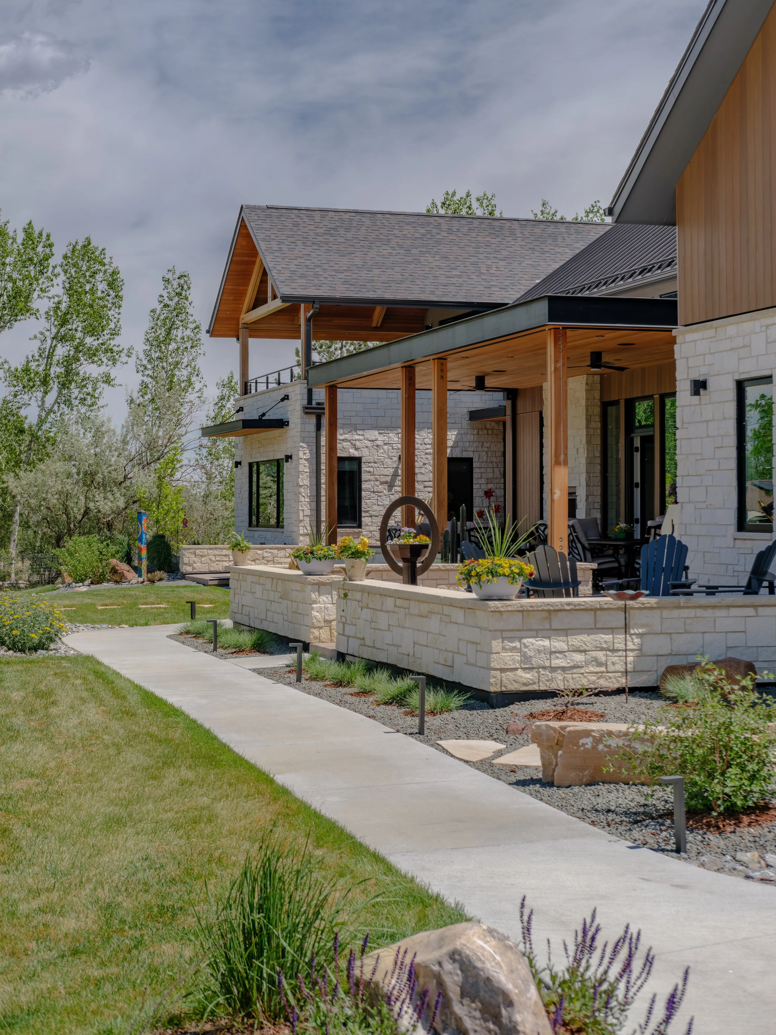 A modern house with a covered porch, wooden accents, large windows, and a well-maintained garden with plants and flowers. The sky is partly cloudy.