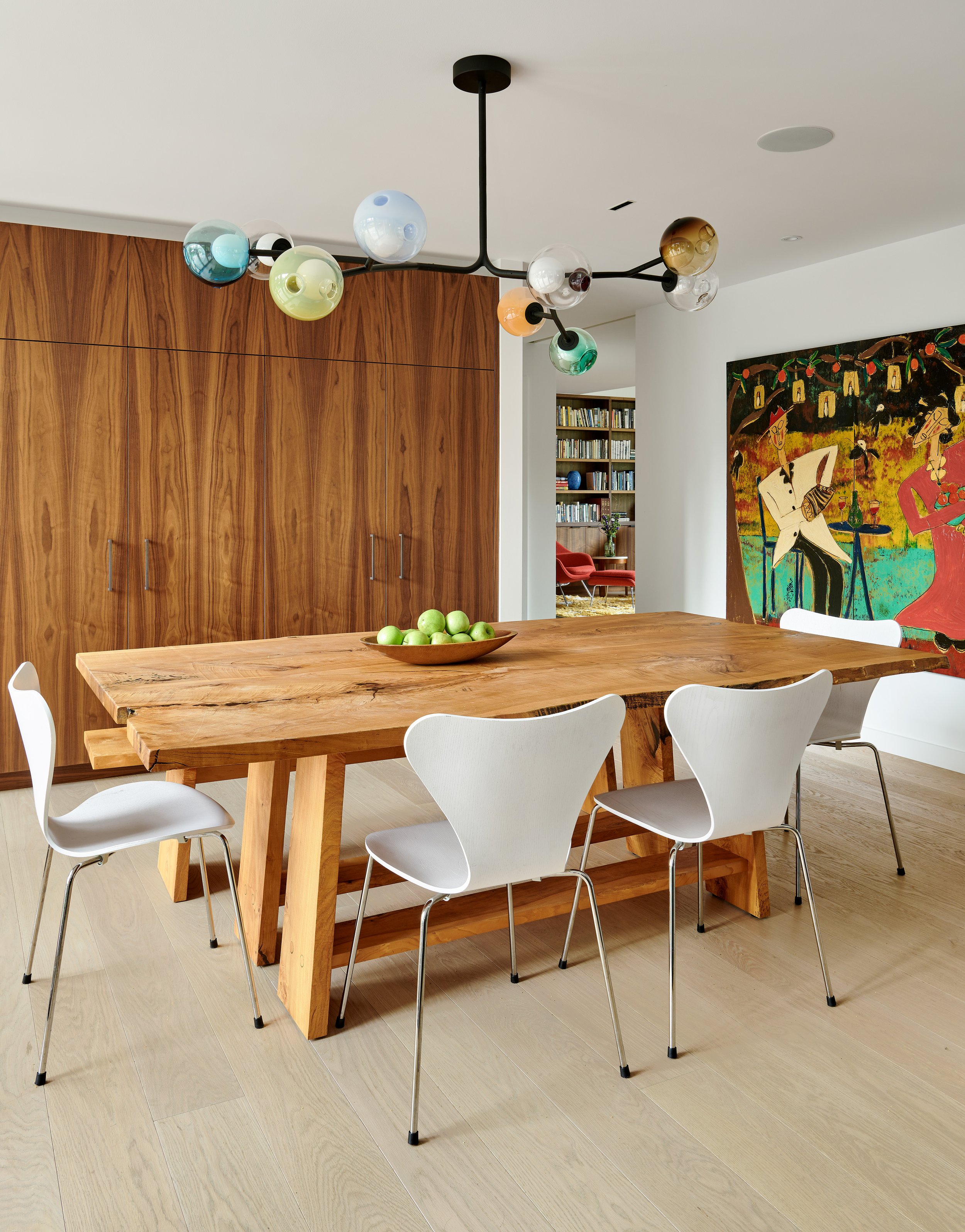 Modern dining room with a large wooden table, white chairs, overhead black chandelier with colorful glass globes, wooden cabinets, a colorful artwork on the wall, and a bookshelf in the background.