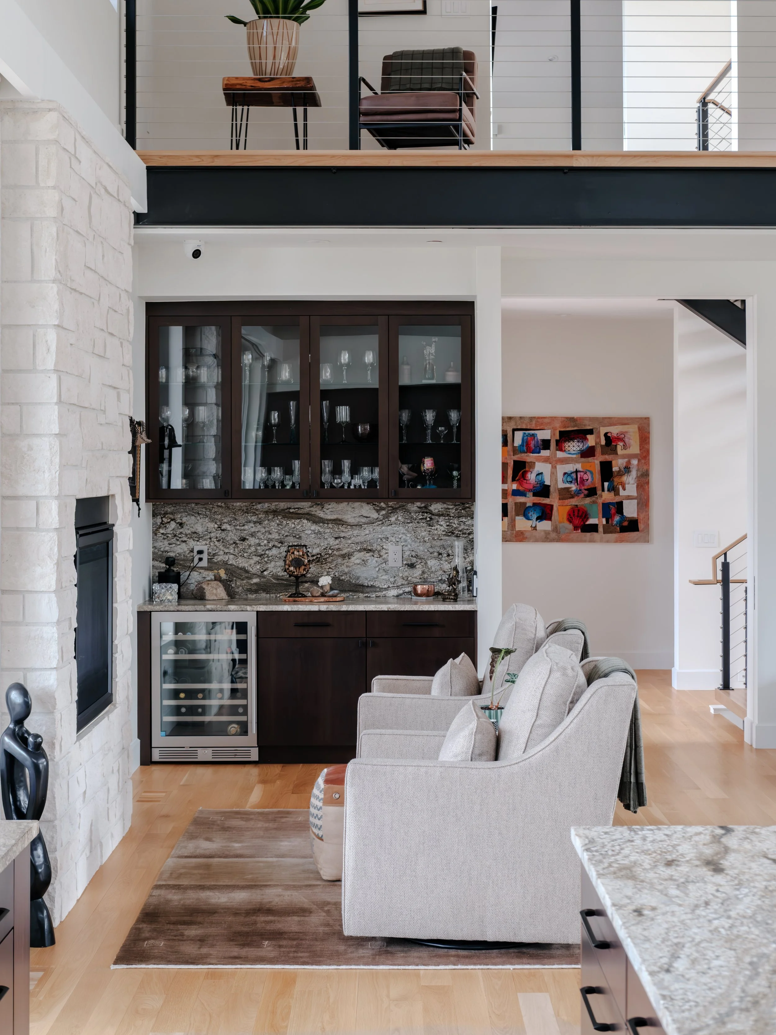 Living room with beige armchairs, a stone fireplace, a rug, and a kitchen counter with a granite top.