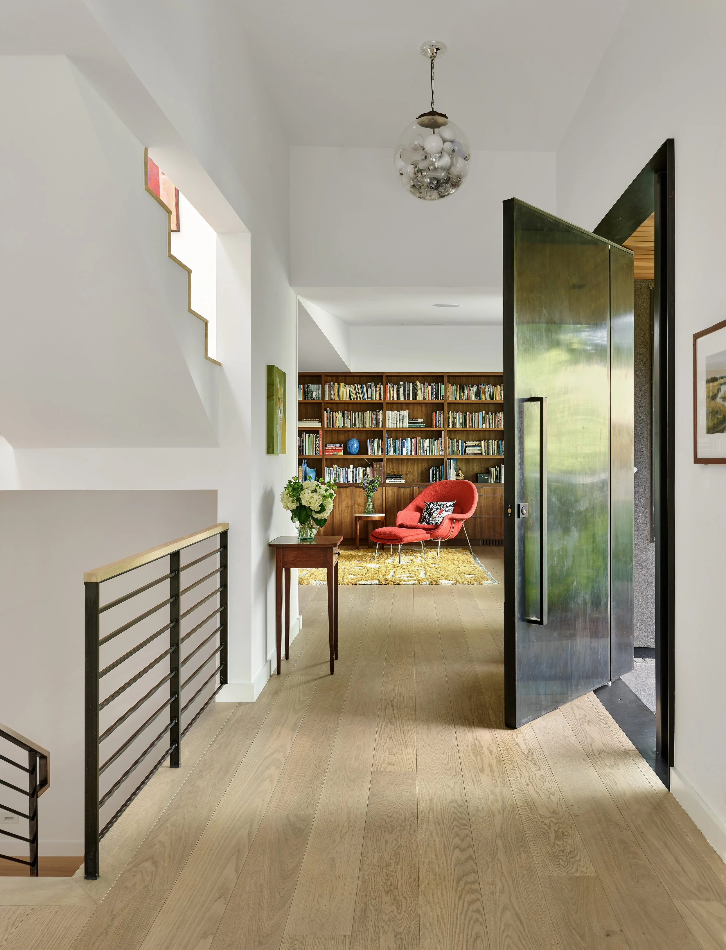Interior view of a modern home with a wooden floor, a bookshelf filled with books, a red lounge chair with a pillow, a side table with flowers, and a unique ceiling light fixture.