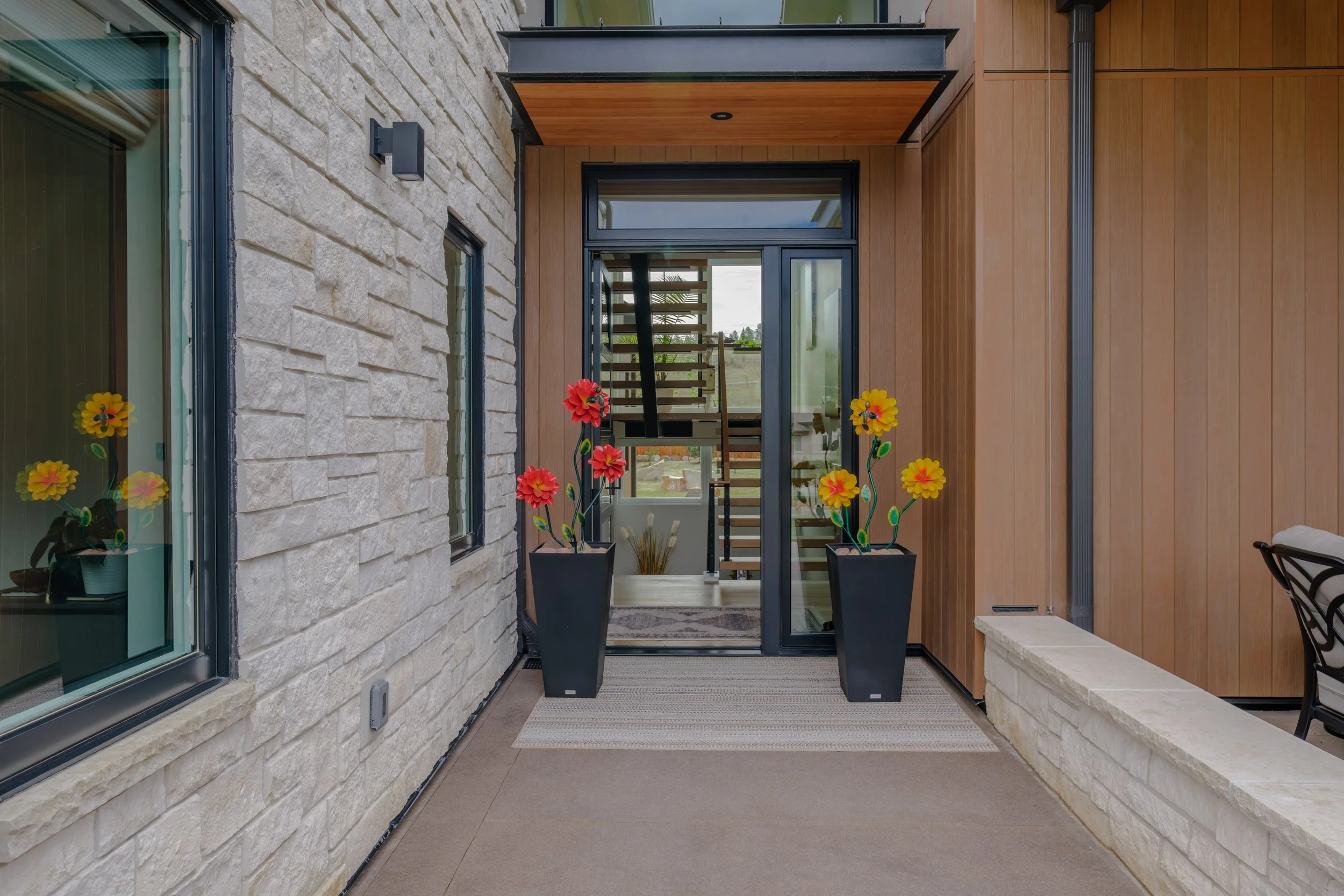 View of a small balcony or porch area with a glass door, two large potted flowers, and a staircase inside the building visible through the glass door. The walls are made of stone and wood, with a light-colored stone wall on the left and vertical wood