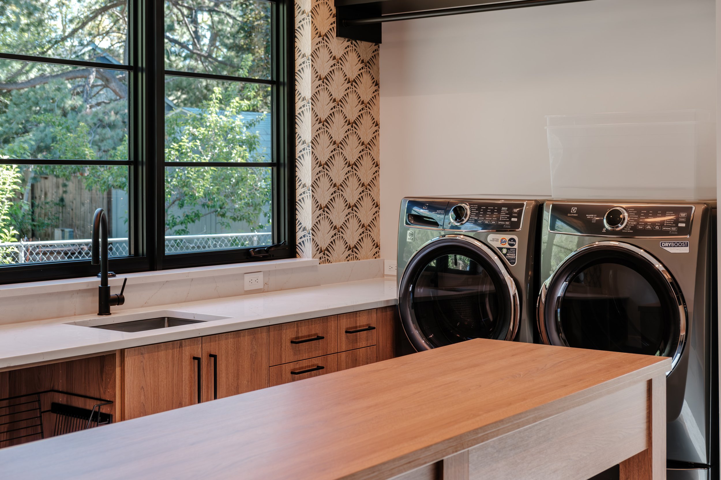 A modern laundry room with a large window, wooden cabinets, a white countertop, and a front-loading washing machine and dryer.