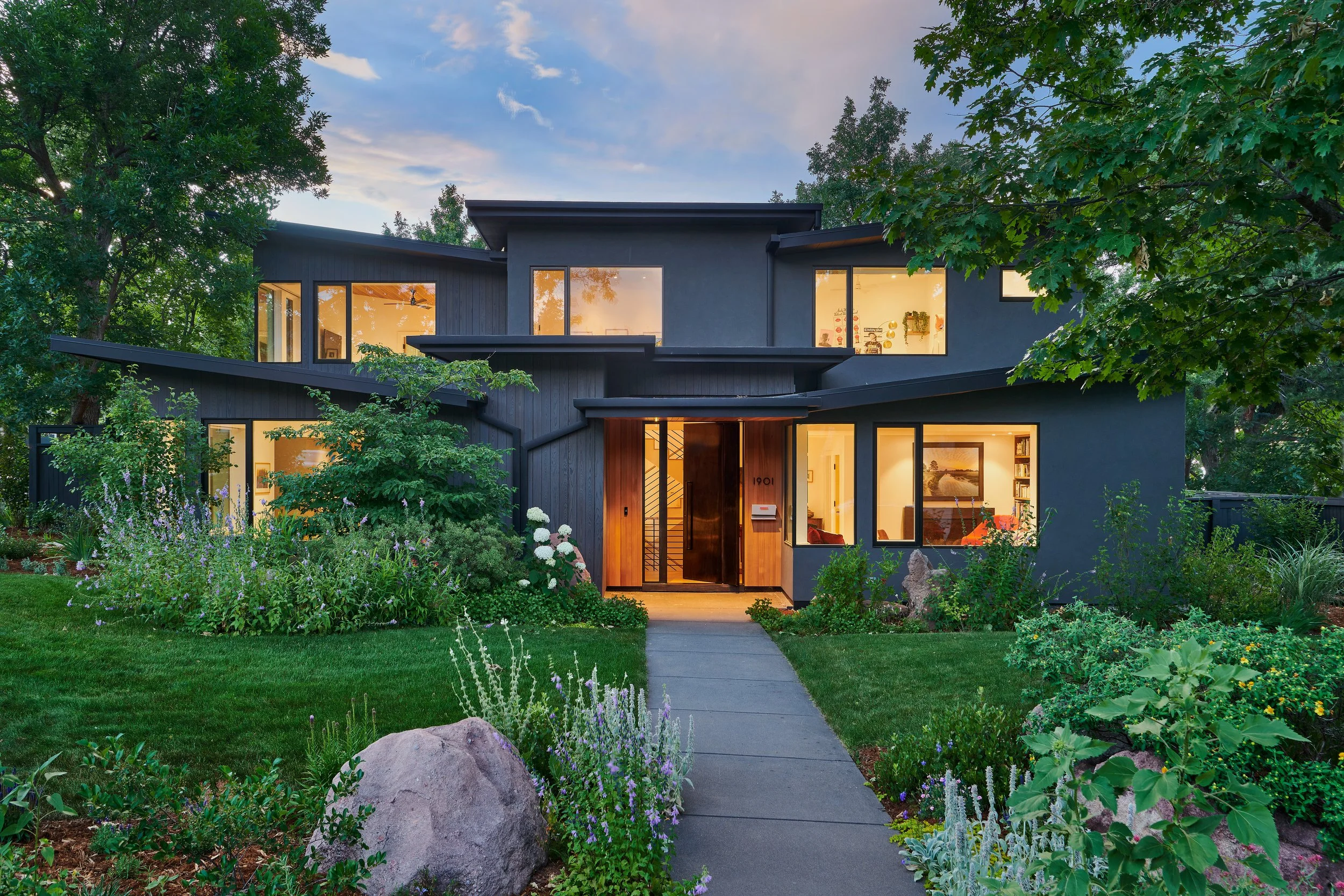 Modern two-story house surrounded by lush green garden, illuminated from inside at dusk.