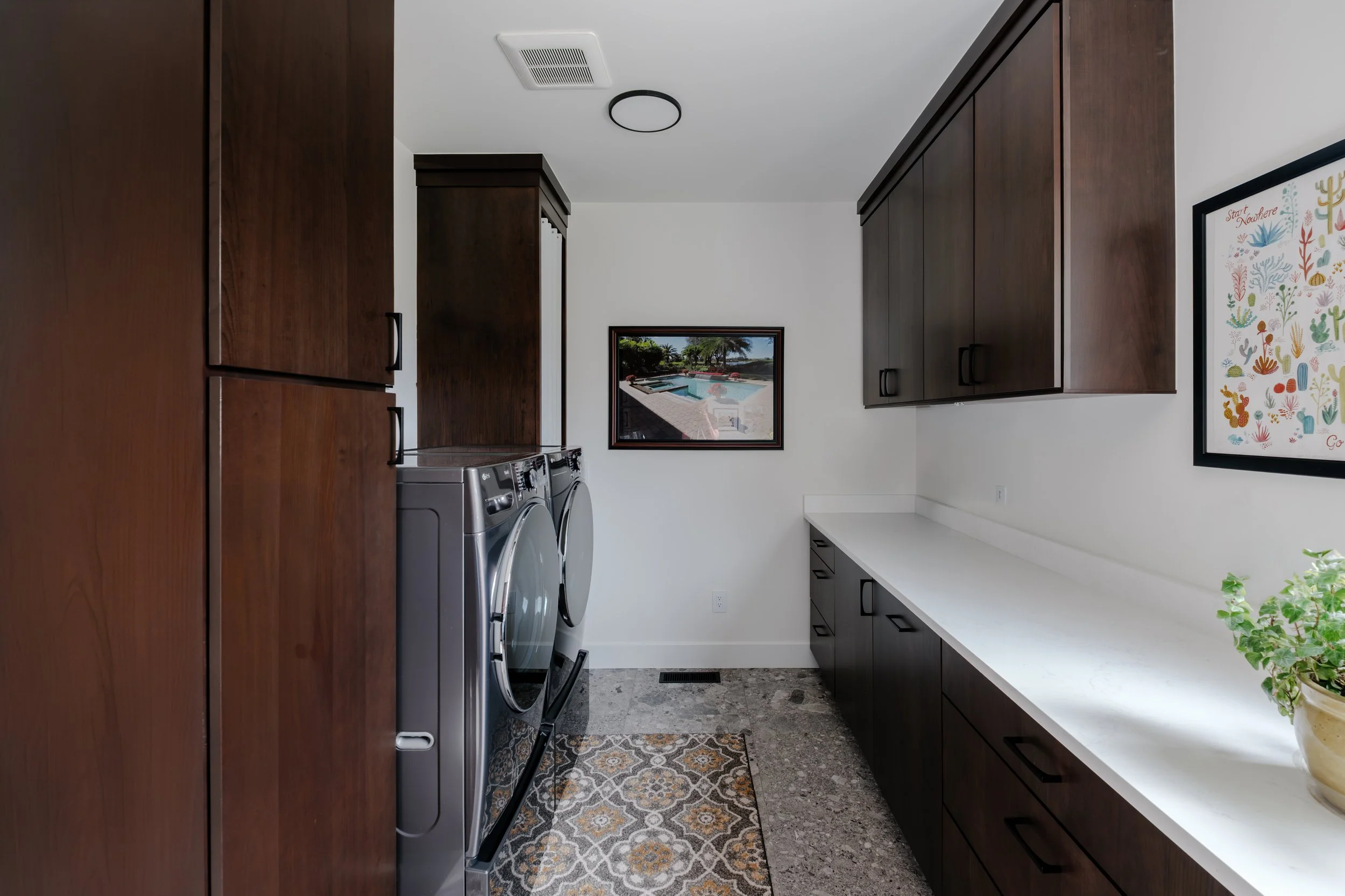 A laundry room featuring dark wooden cabinets, a white countertop, a front-loading washer and dryer, framed artwork on the wall, and a patterned rug on the floor.