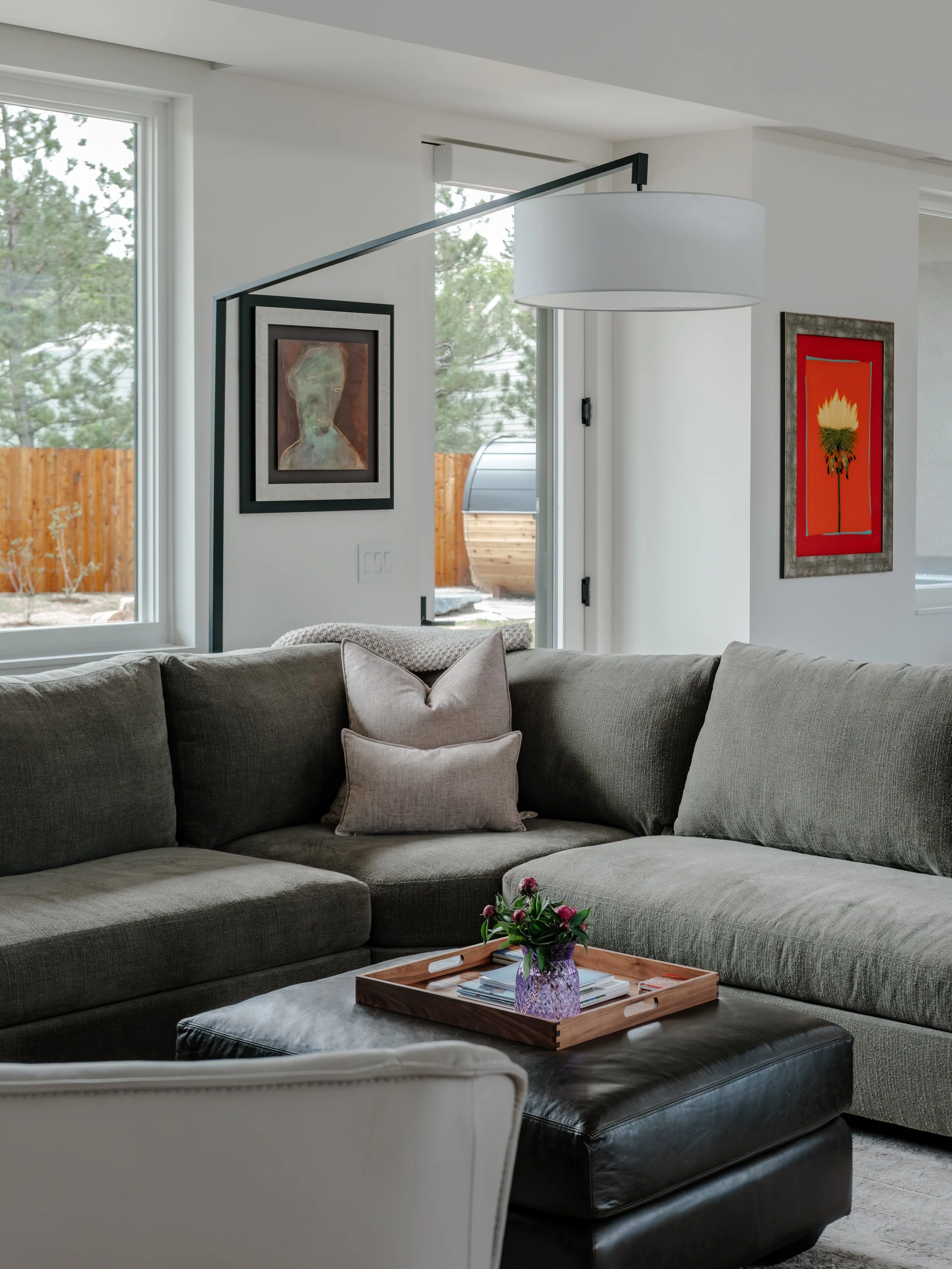 Living room with a large gray sectional sofa, a white leather chair, a coffee table with a tray and flowers, and artwork on the walls. Large windows show trees outside.