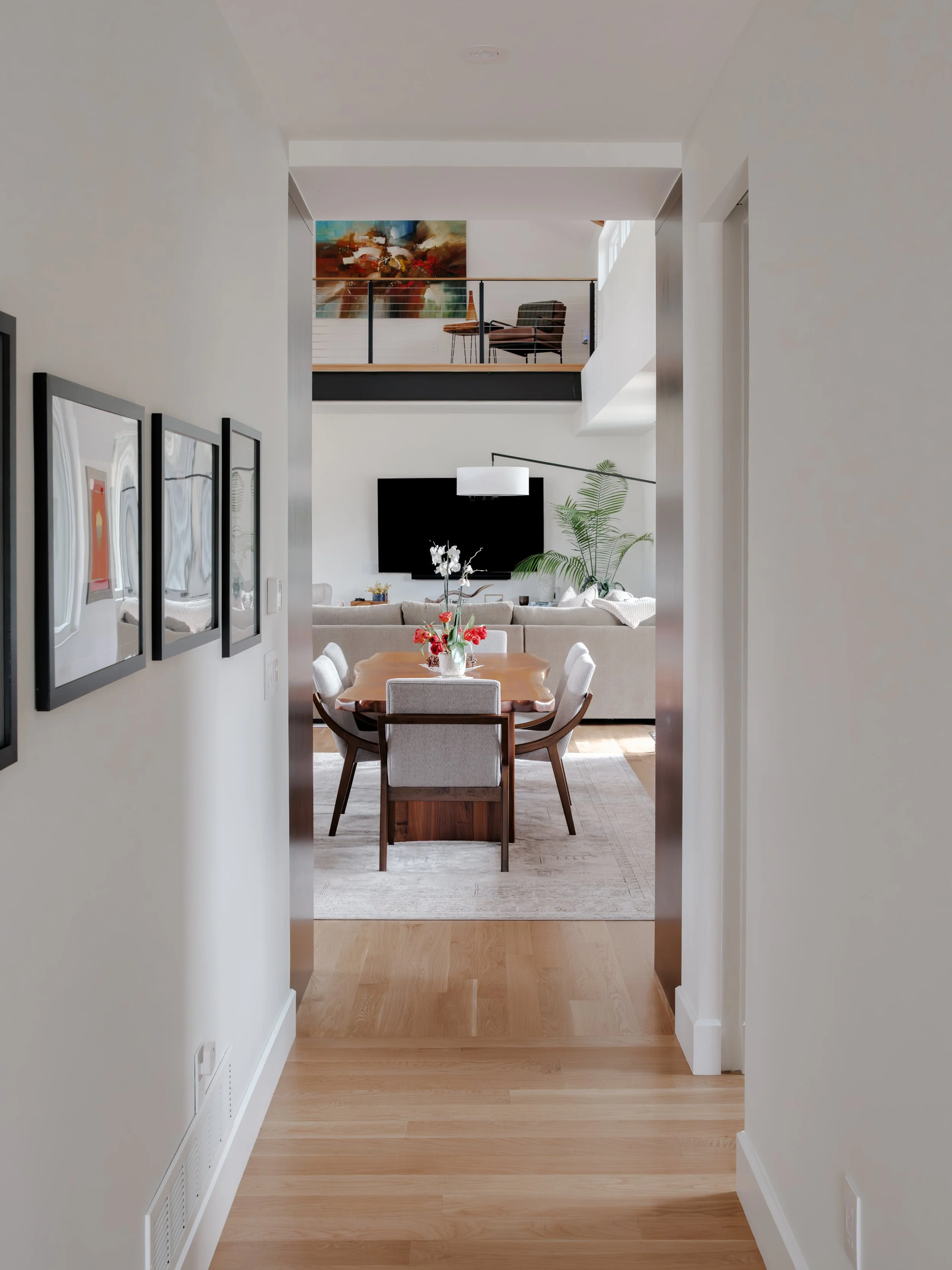 View of a living room from a hallway, with a dining table and chairs in the foreground, a large flat-screen TV, and a balcony with chairs and a colorful abstract painting on the wall in the background.