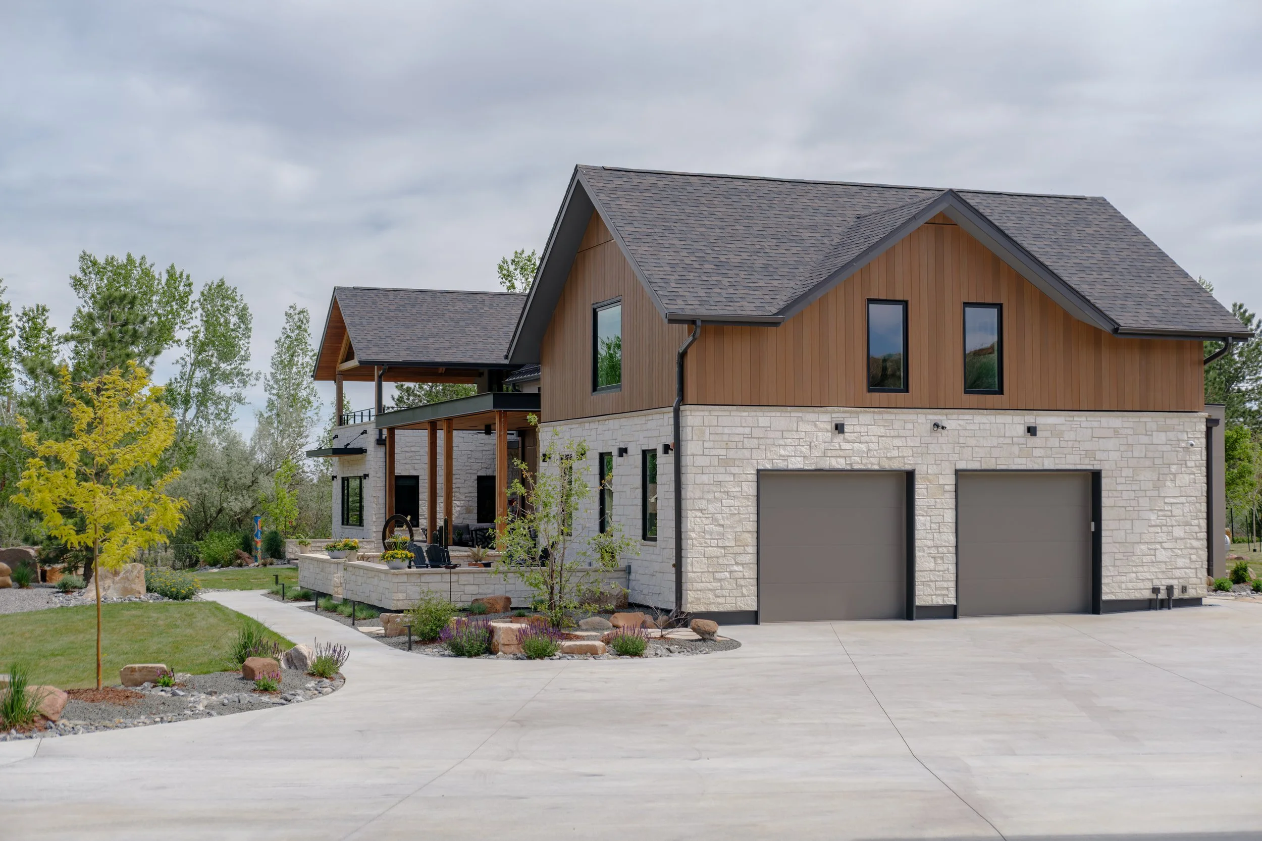 A modern two-story house with stone and wooden exterior, surrounded by a landscaped yard with a curved sidewalk and trees.