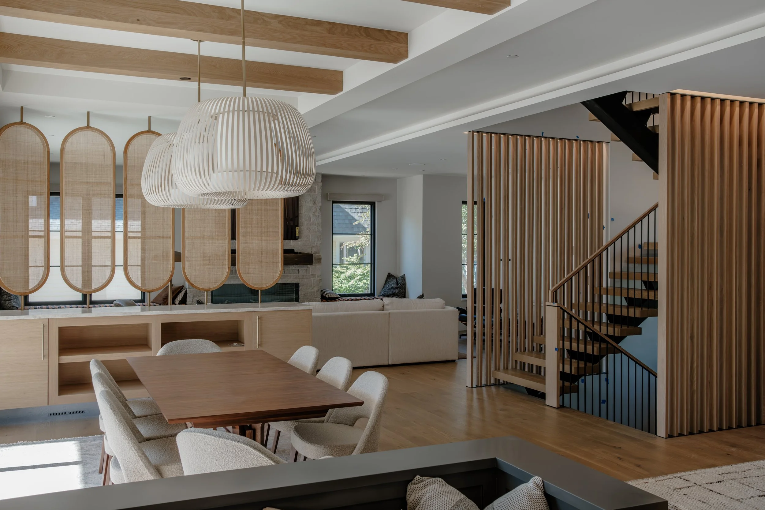 Interior view of a modern living space featuring a dining area with a wooden table and beige chairs, a cozy sitting area with a white sofa and armchair, a staircase with wooden and black elements, vertical wooden slats, large windows allowing natural