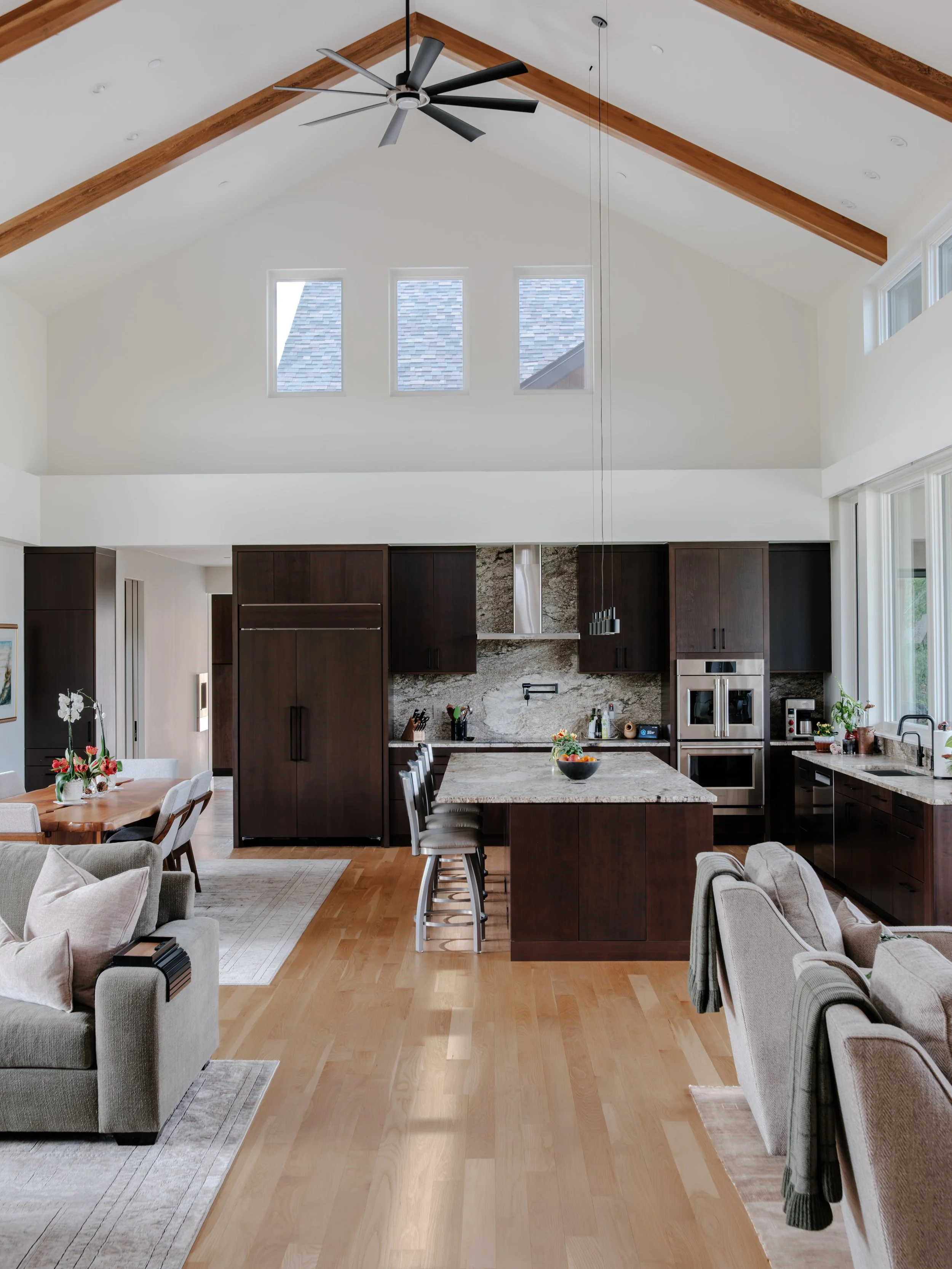 Open-concept living area with kitchen and dining space, high vaulted ceiling, wooden beams, modern ceiling fan, three rectangular windows, dark wood cabinets, marble backsplash in the kitchen, light wood flooring, beige sofas, and a dining table with
