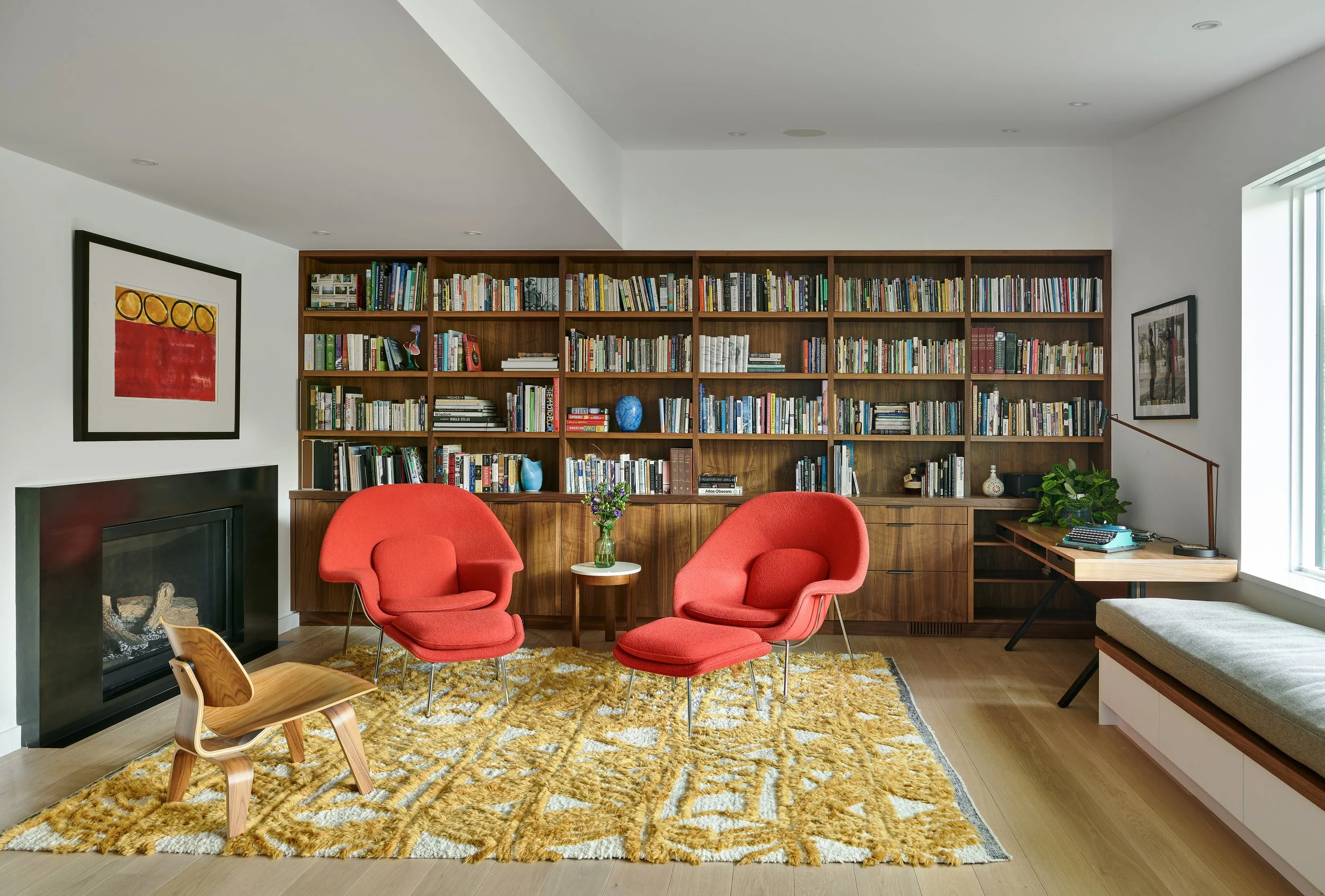 A cozy living room with a large wooden bookshelf filled with books, a fireplace, and vibrant red armchairs around a small white table with purple flowers. There is a textured yellow rug underneath the chairs and a window allowing natural light.
