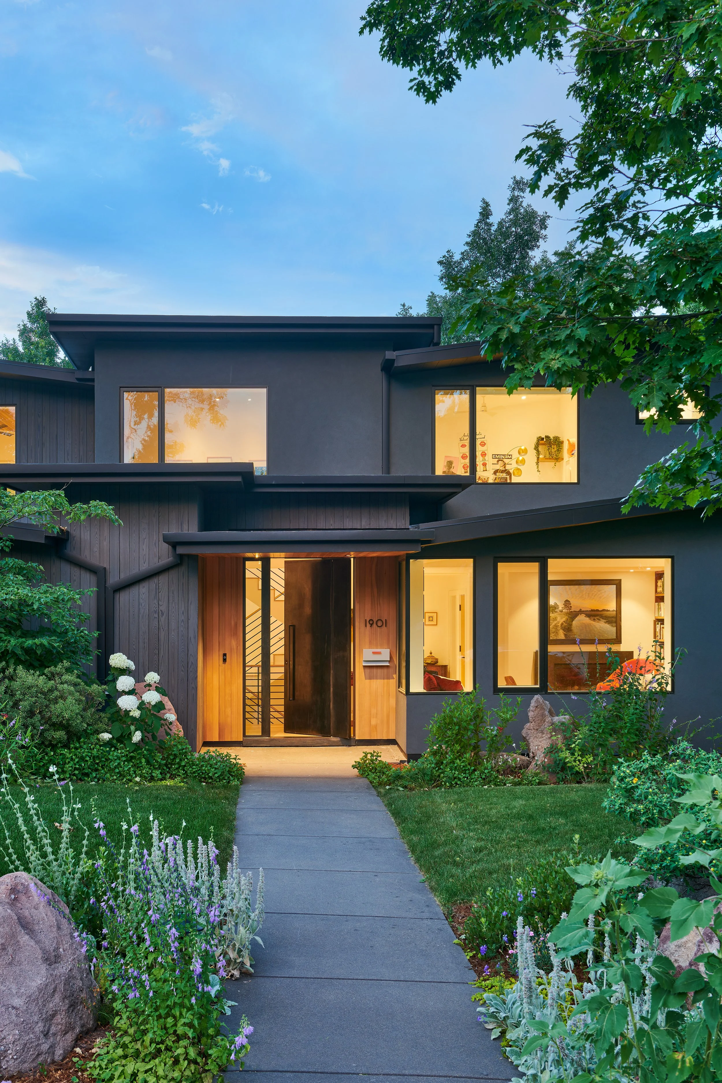 Modern house with large windows, wooden and dark gray exterior, surrounded by lush green garden with flowers, plants, and rocks, walkway leading to front door, evening lighting inside illuminating the house.