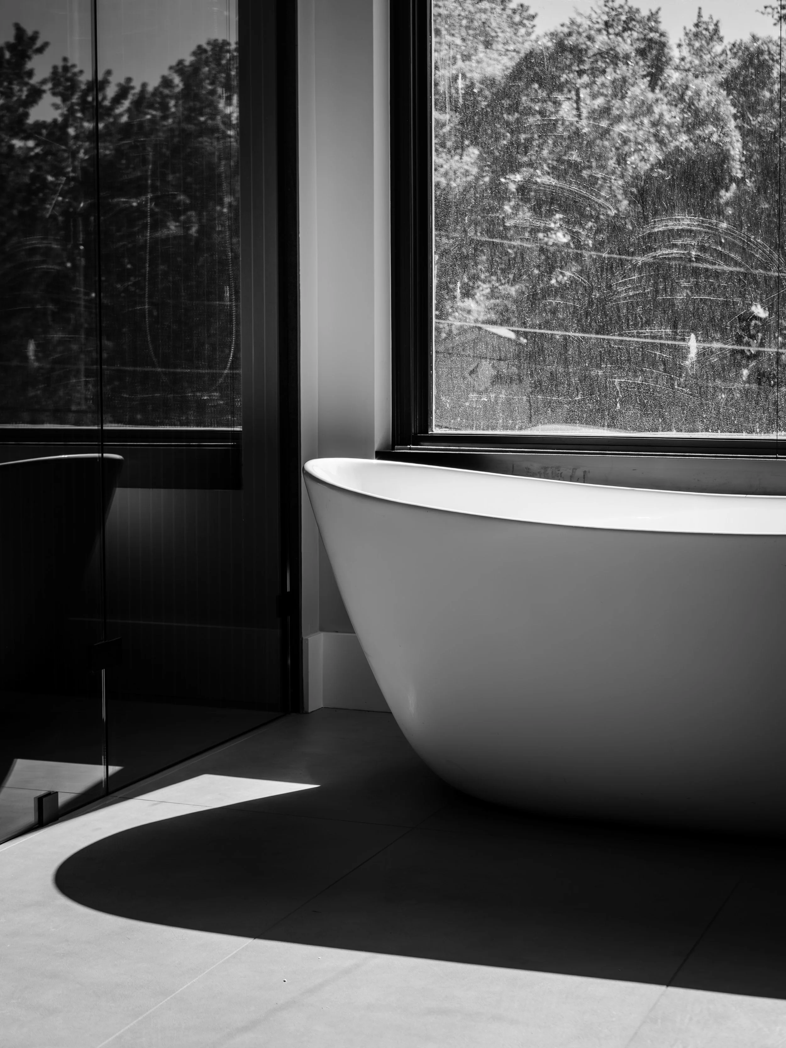 Black and white photo of a modern bathtub next to a large window with a view of trees outside. The shadow of the bathtub is cast on the floor.
