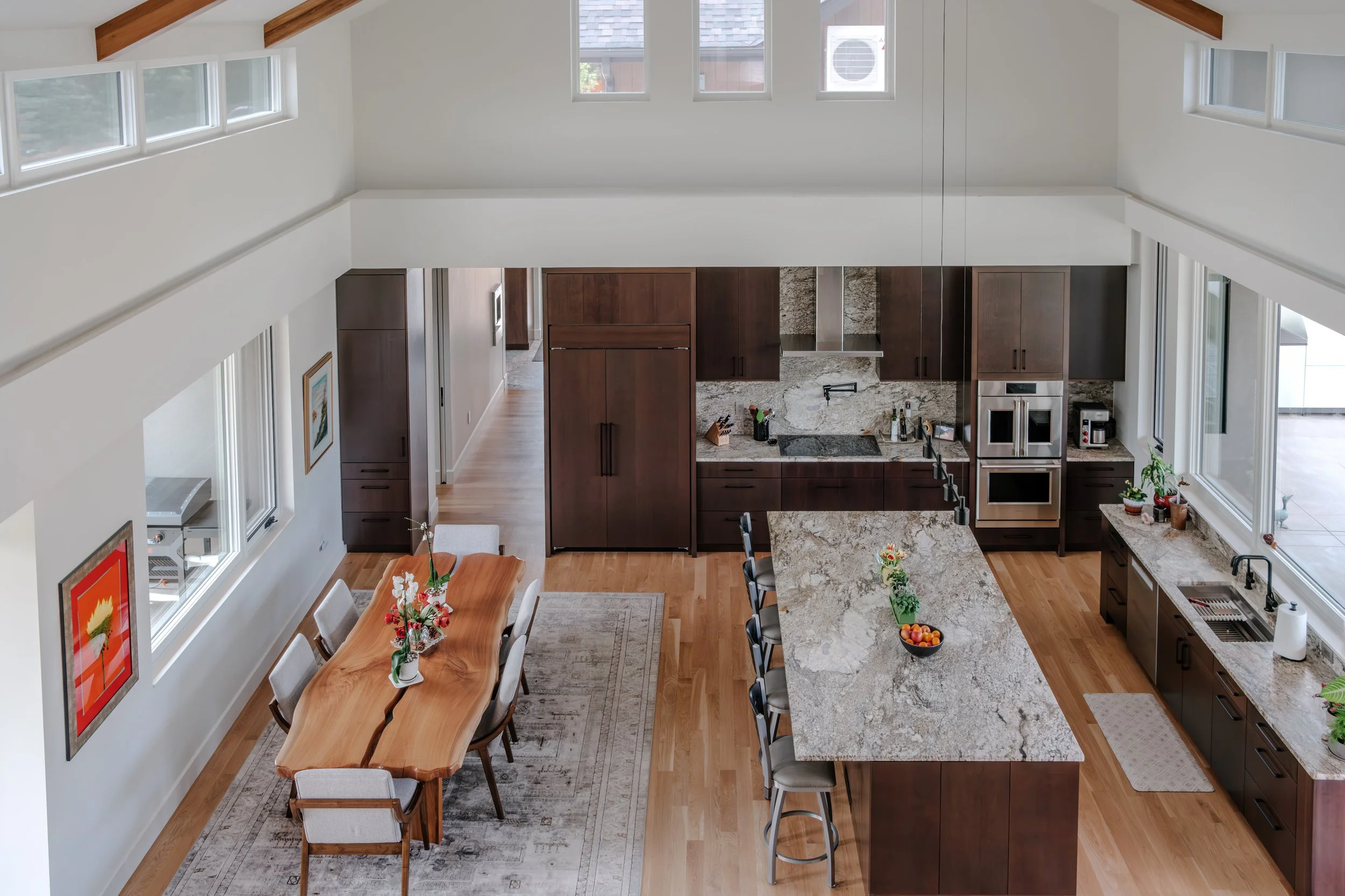 Open-concept kitchen and dining area with vaulted ceiling, wooden beams, large windows, dark wood cabinets, granite countertops, and a dining table with chairs and flower arrangements.