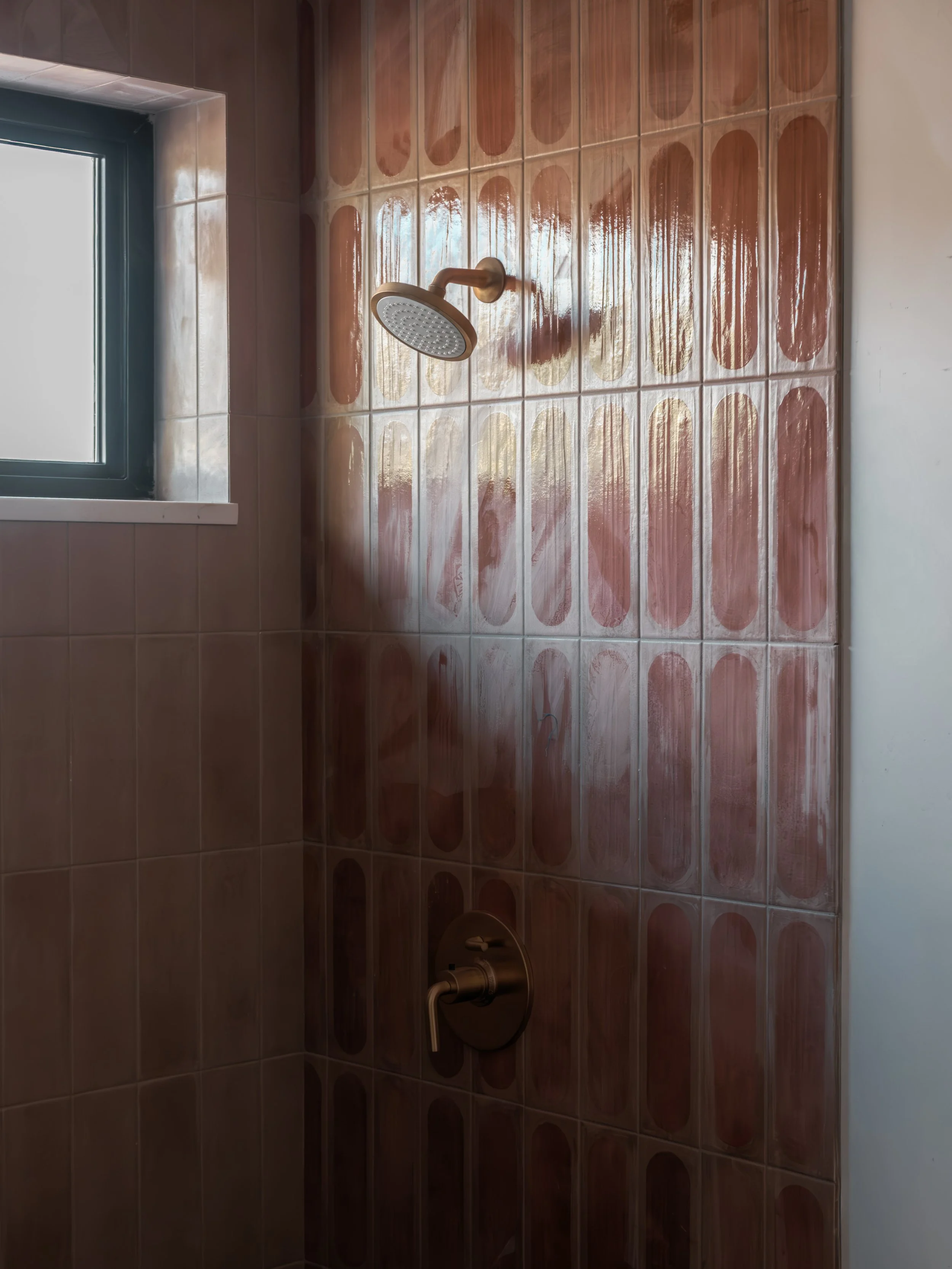 A shower area with brown and beige oval patterned tiles, a mounted shower head, and a window on the upper left side.