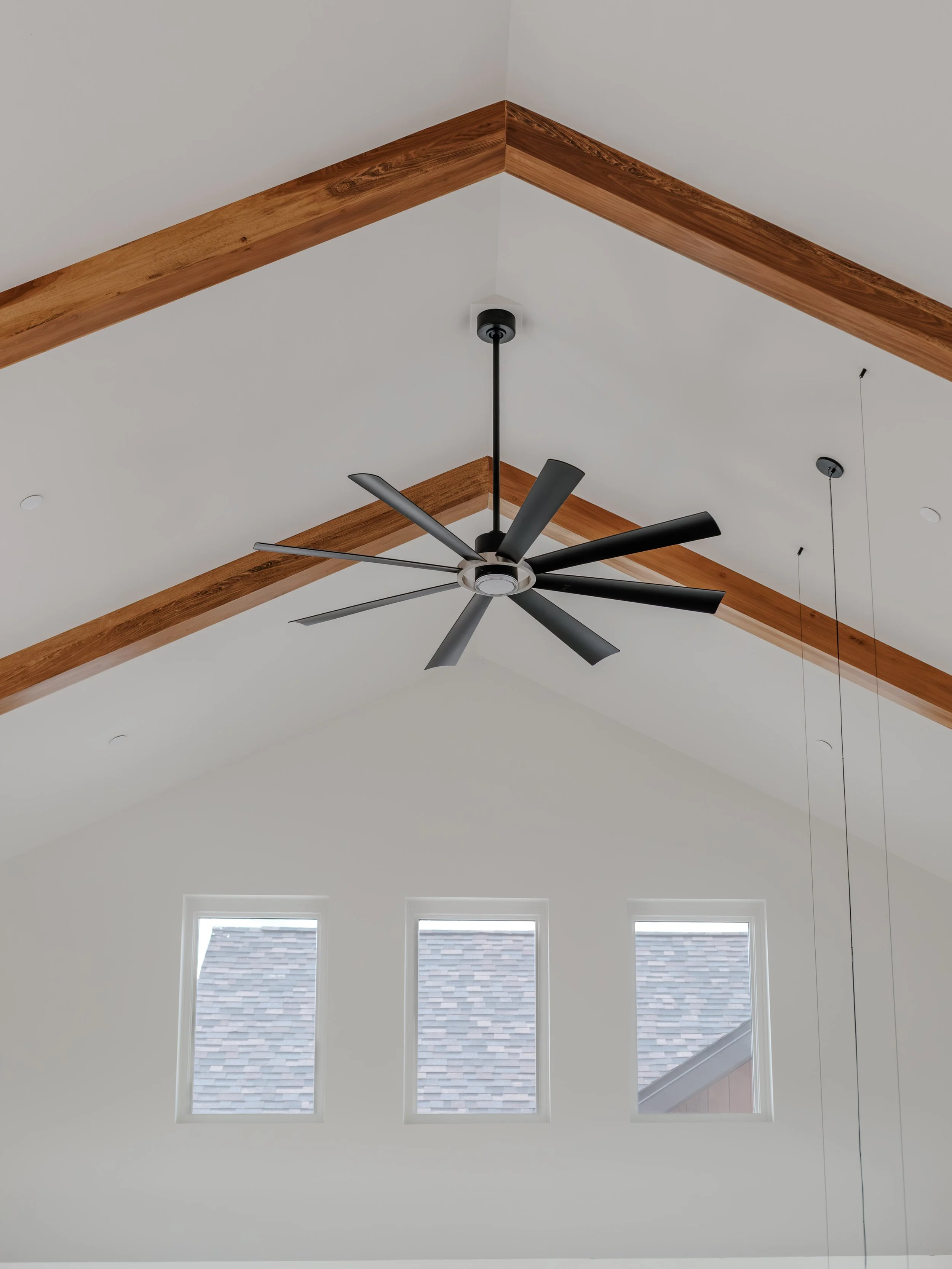 View of a modern ceiling with three tall rectangular windows, a black ceiling fan with multiple blades, and wooden beams in a spacious room.