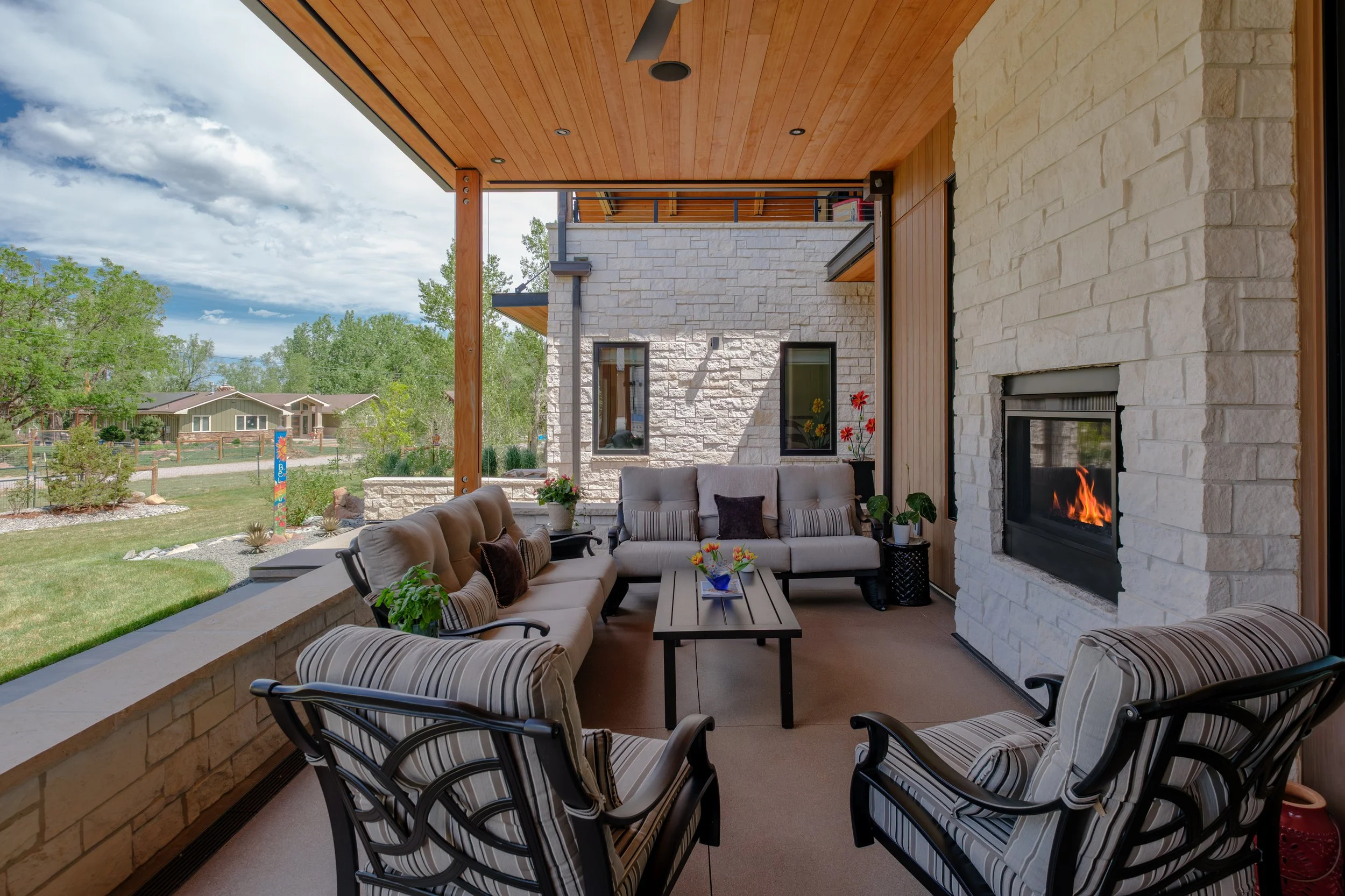 Covered outdoor patio with comfortable seating, a fireplace, and a view of a yard with trees and houses under a cloudy sky.