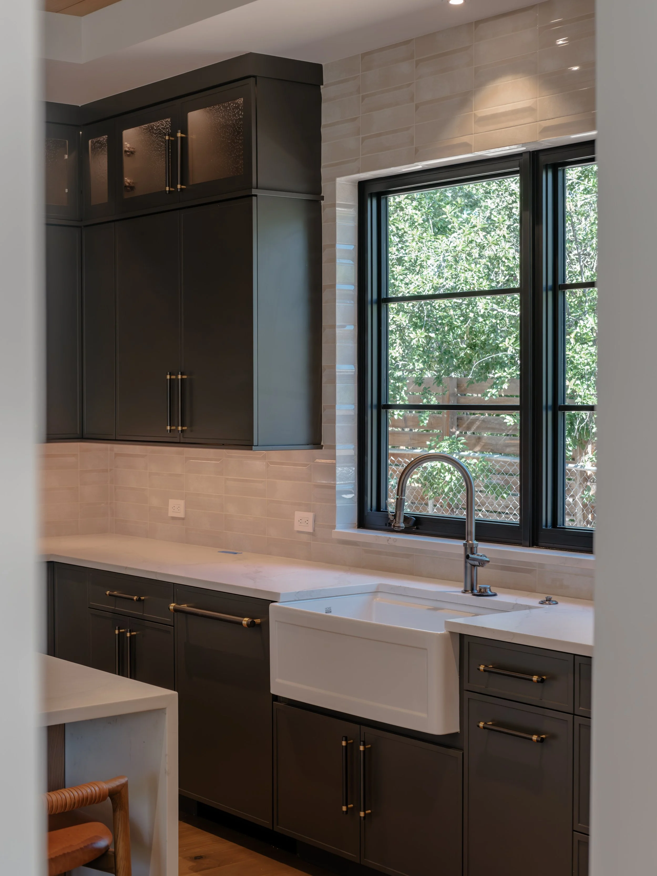 A modern kitchen with dark gray cabinets, a white farmhouse sink, and a large window with black framing showing greenery outside.