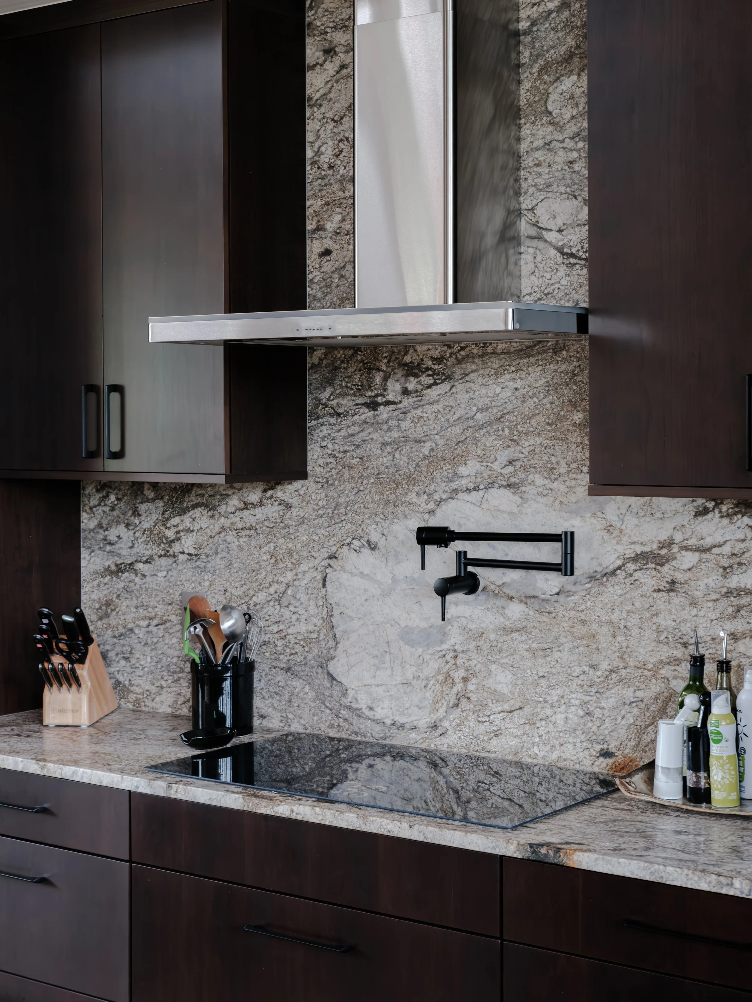 Kitchen counter with dark wooden cabinets, a black utensil holder, and bottles near a beige and brown granite countertop and backsplash, with a black faucet and stovetop.