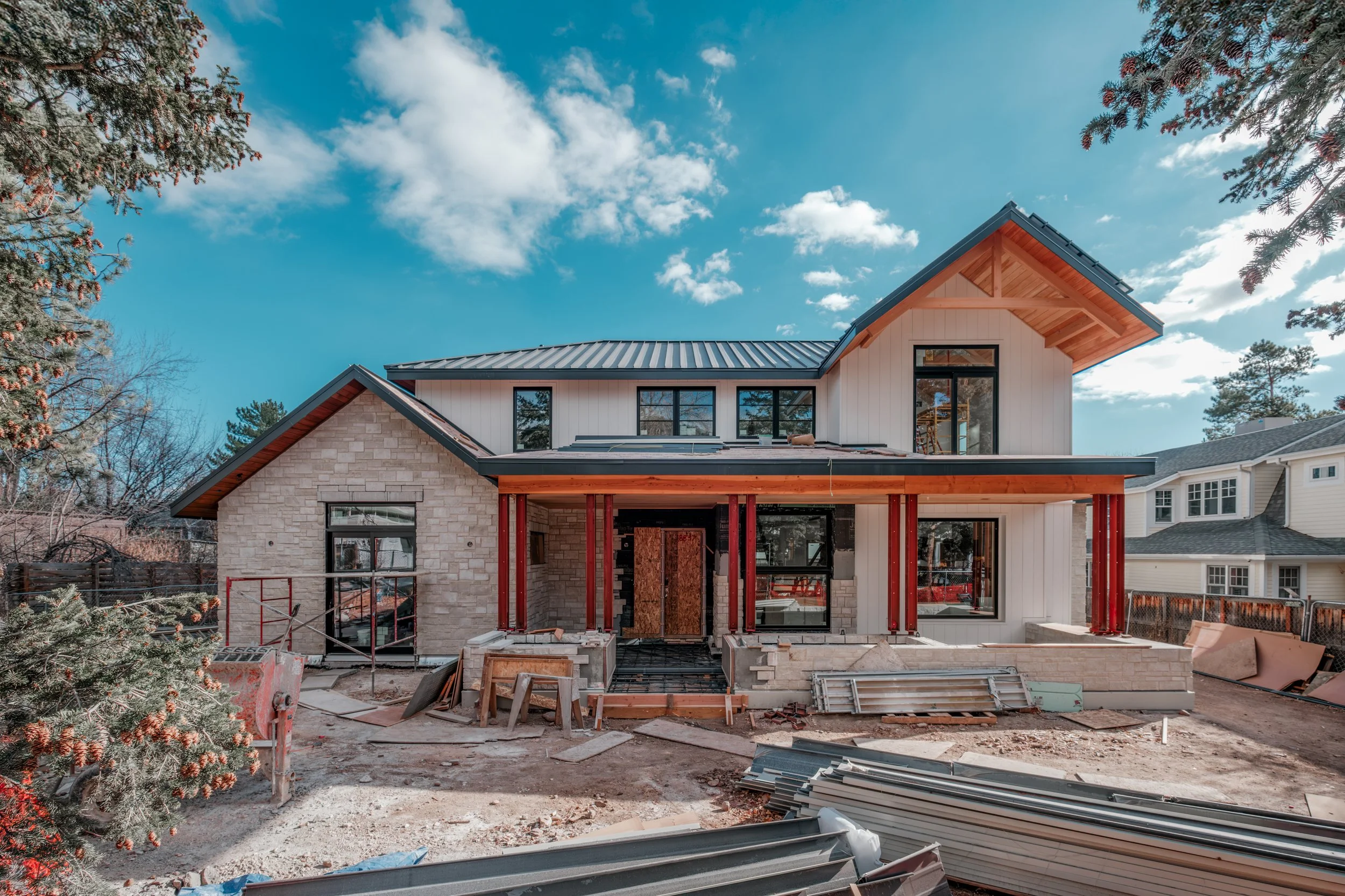 A two-story house under construction with scaffolding and building materials in the front yard, surrounded by tall pine trees and a partly cloudy blue sky.