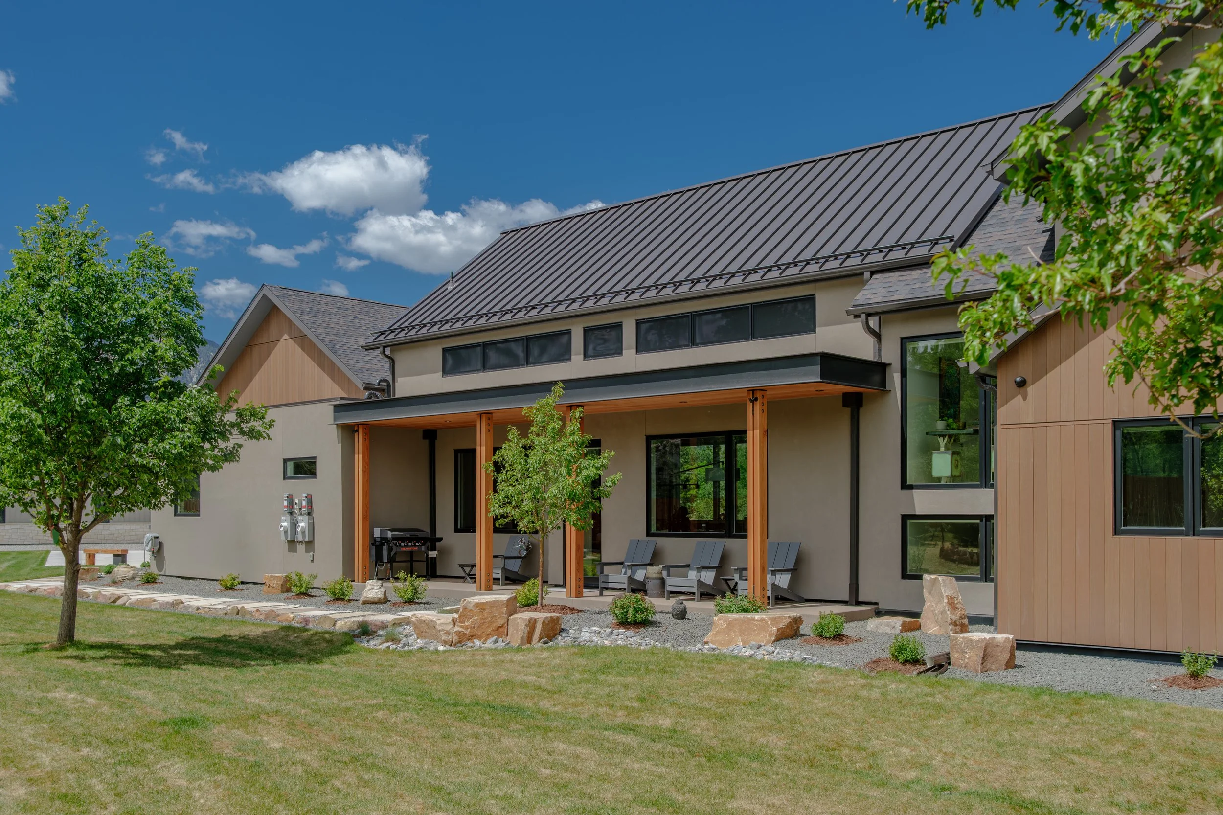 Modern house with a metal roof, large windows, and a small patio area with outdoor chairs and a barbecue, surrounded by green trees and a lawn, under a bright blue sky with some clouds.