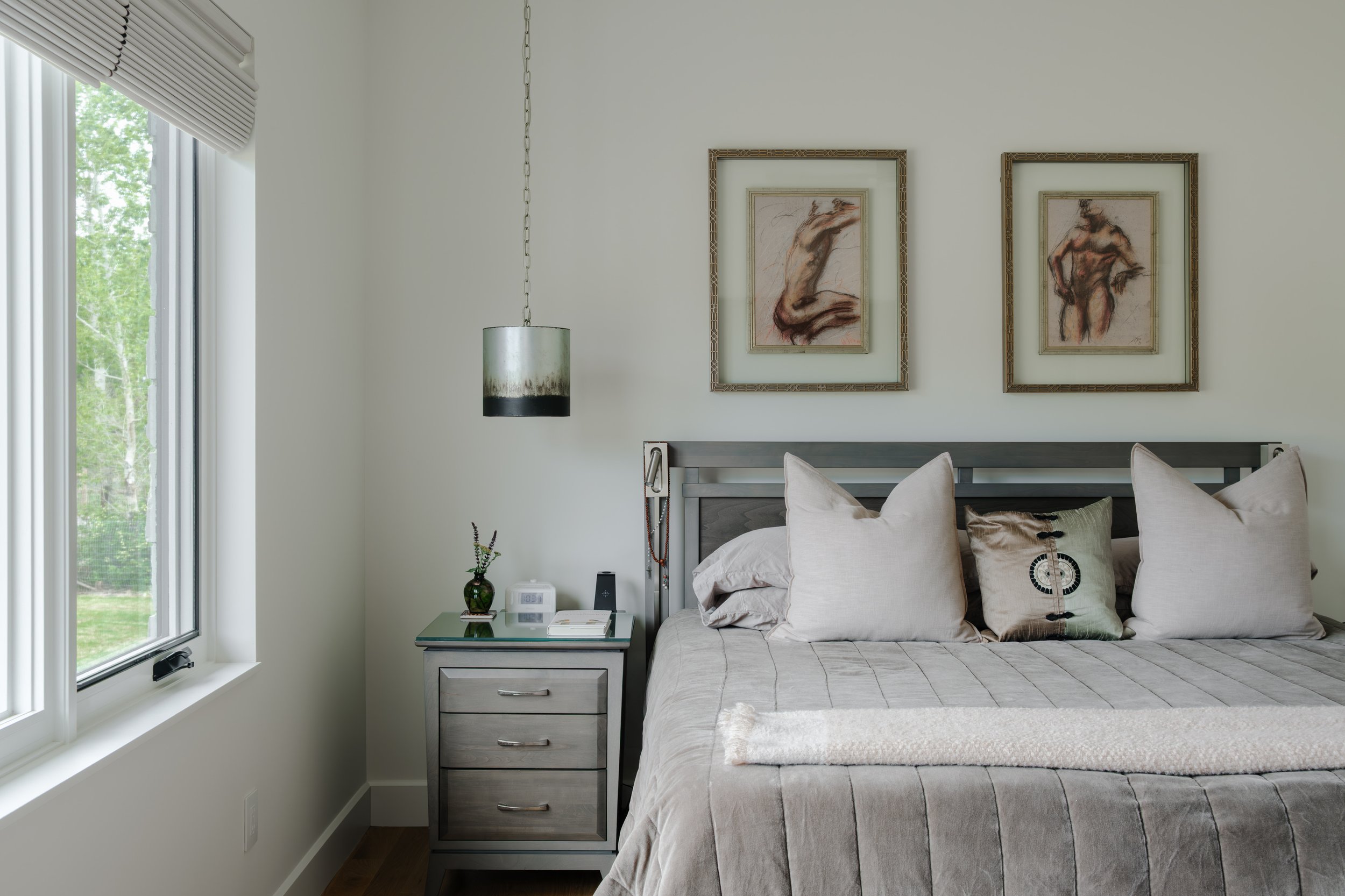 A bedroom with a large window on the left, a nightstand with a small green vase, a clock, and some books. The bed has beige pillows and a beige blanket, with two framed artistic sketches of nude figures hanging on the white wall above the headboard. 