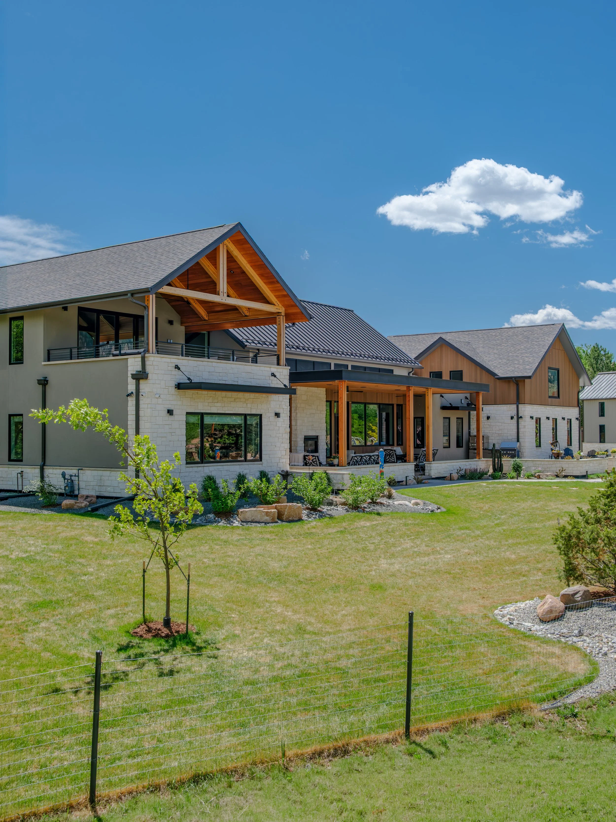 Modern residential building with a landscaped yard, young trees, metal fences, and a partly cloudy blue sky.
