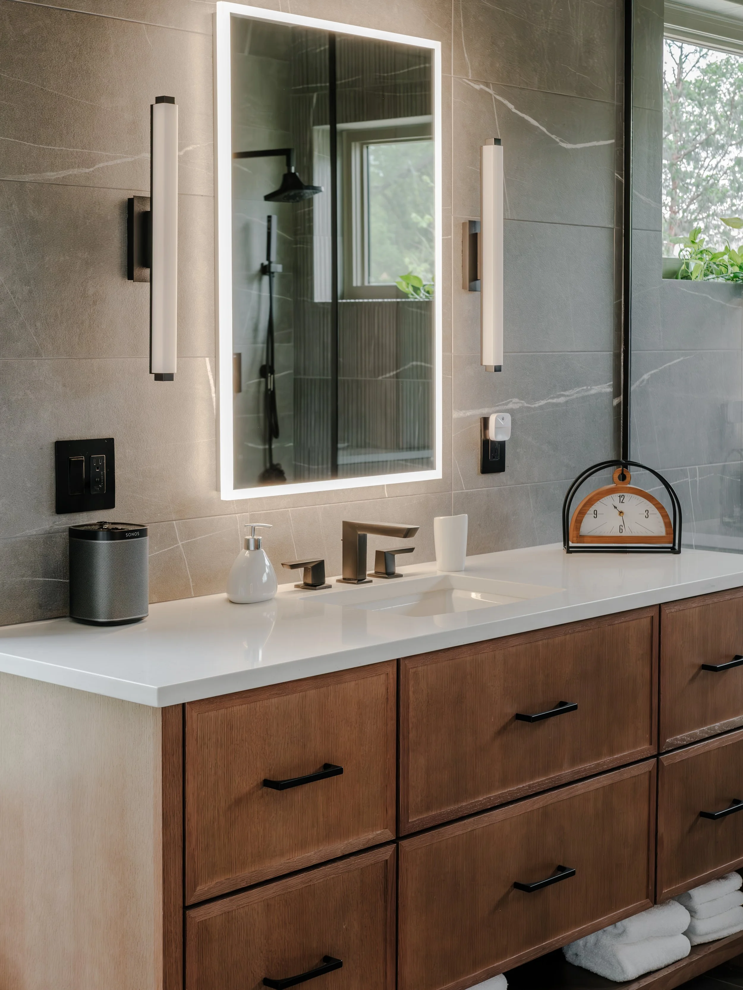 Modern bathroom vanity with a white countertop, wooden cabinets, and a large illuminated mirror. There are two vertical wall lights, a soap dispenser, a small speaker, a clock, and black faucet fixtures. Towels are neatly stored underneath.
