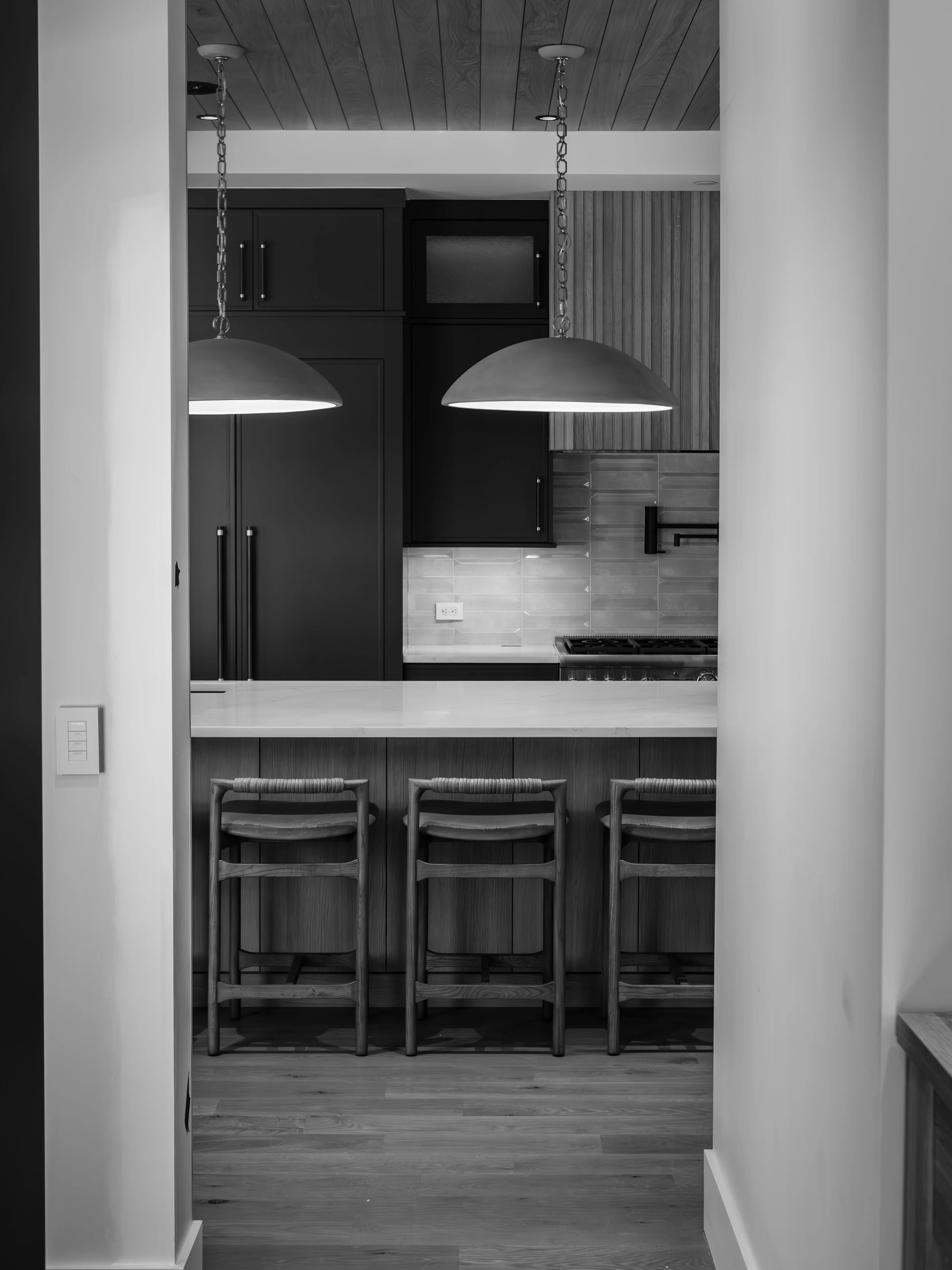 A black-and-white photo of a modern kitchen with a wooden ceiling, two hanging pendant lights over a counter, black cabinets, backsplash, and a stove, with wooden chairs lined up at the counter.