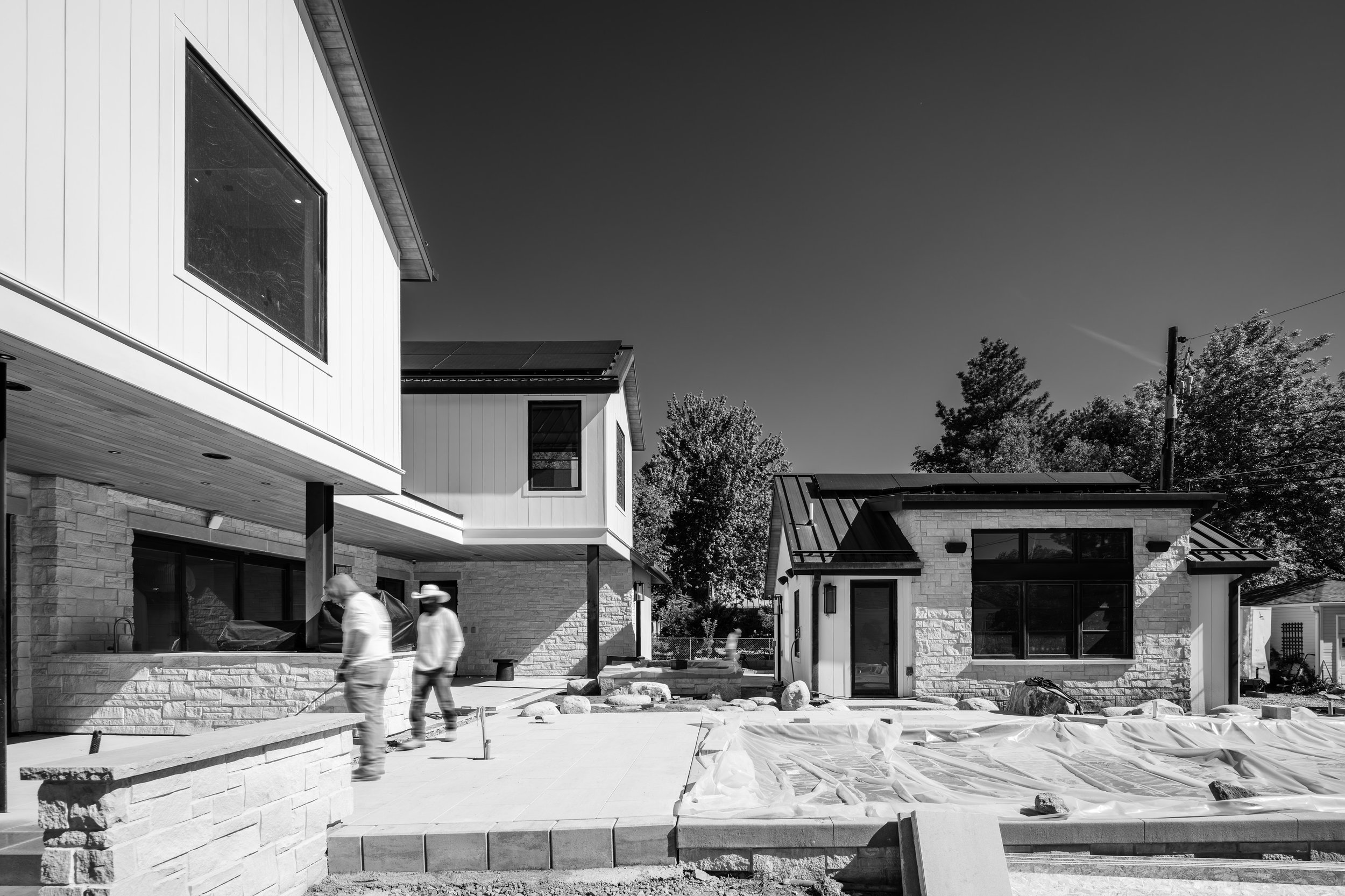 Black and white photo of a modern house under construction with workers in the yard, some with cap and-helmet, near a patio with tiles and a stone wall, several trees in the background.