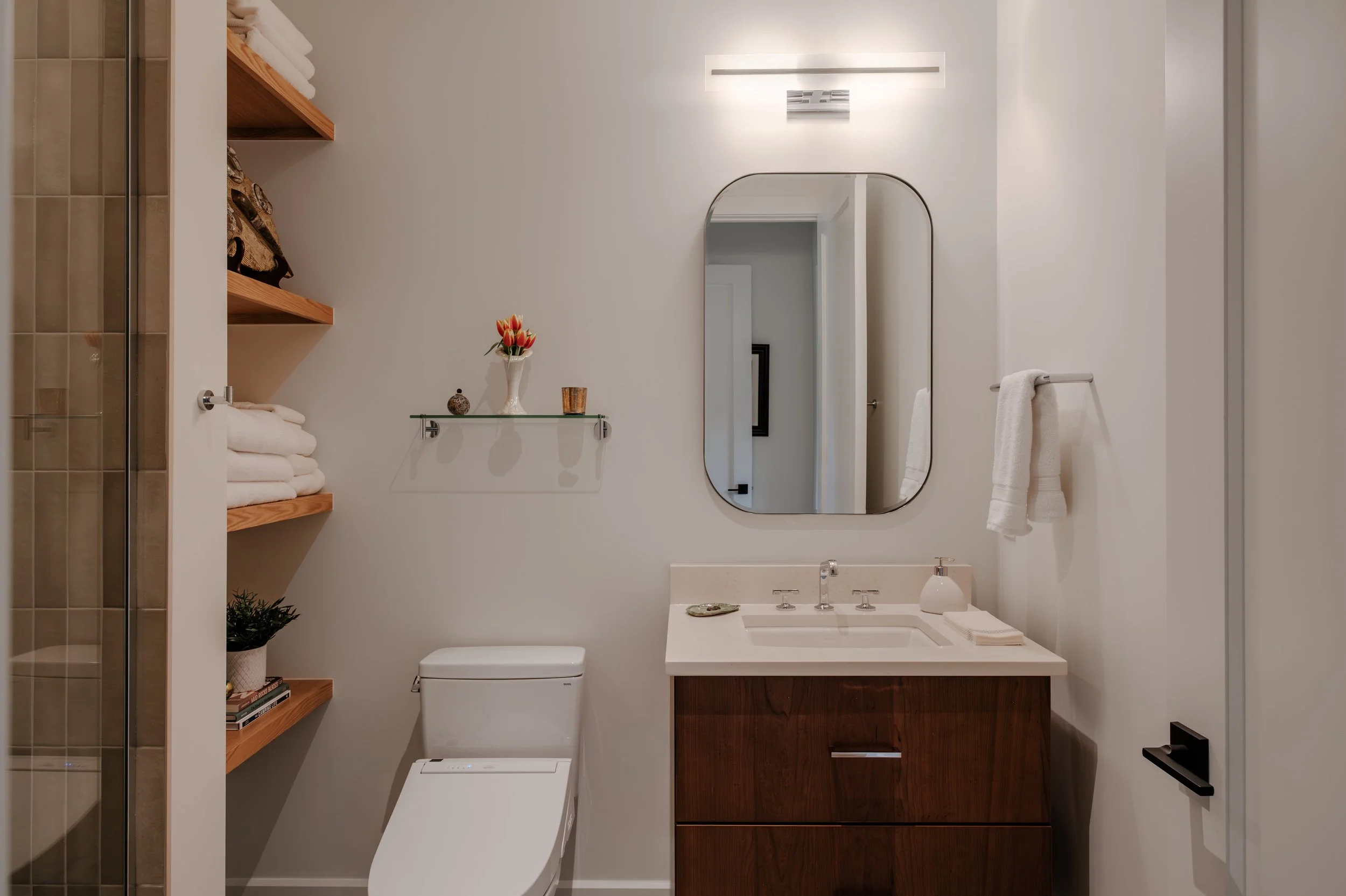 A modern bathroom with a white toilet, a white sink with a wooden cabinet, a large mirror, a wall-mounted light, towels on a rack, and a glass shelf with a flower vase and decorative items.