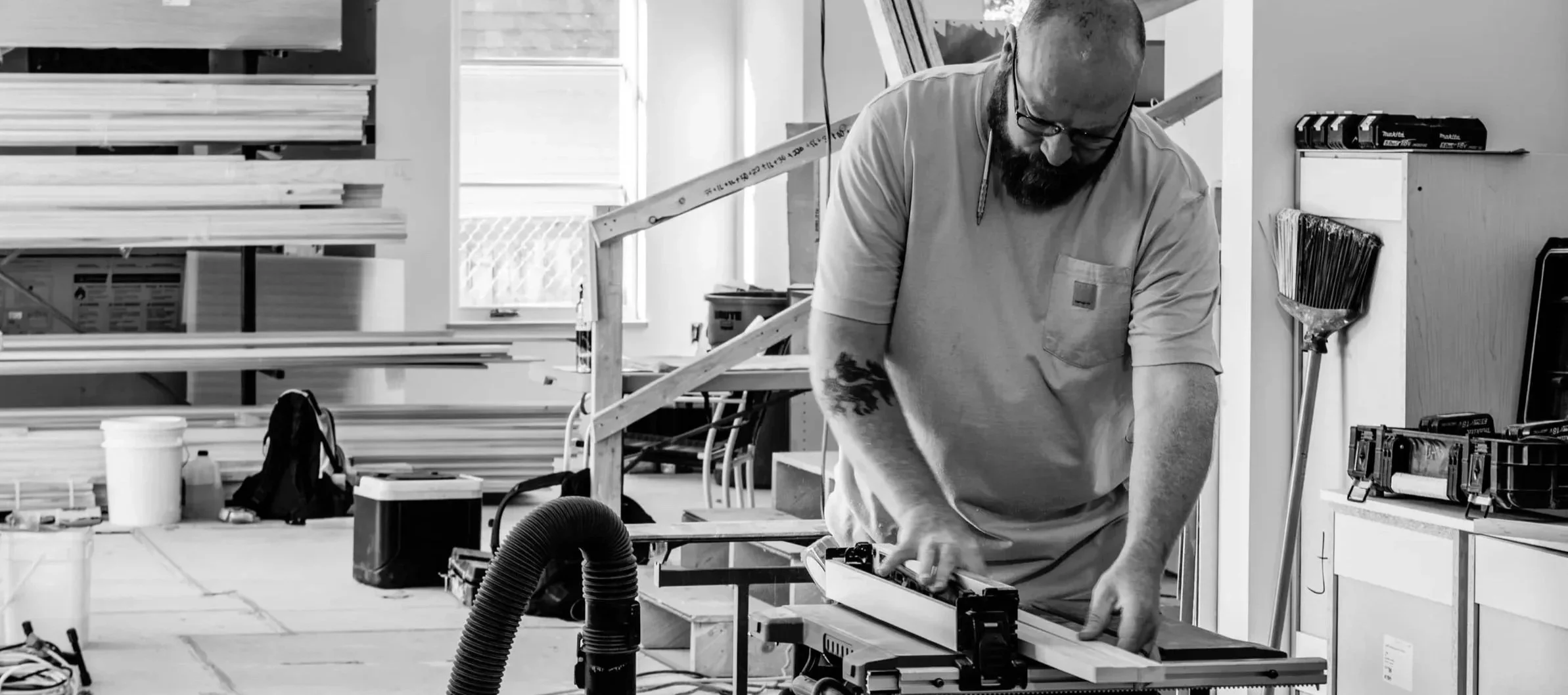A man with a beard, glasses, and tattoos working on a woodworking project in a brightly lit workshop. The workspace is filled with shelves, tools, and materials.