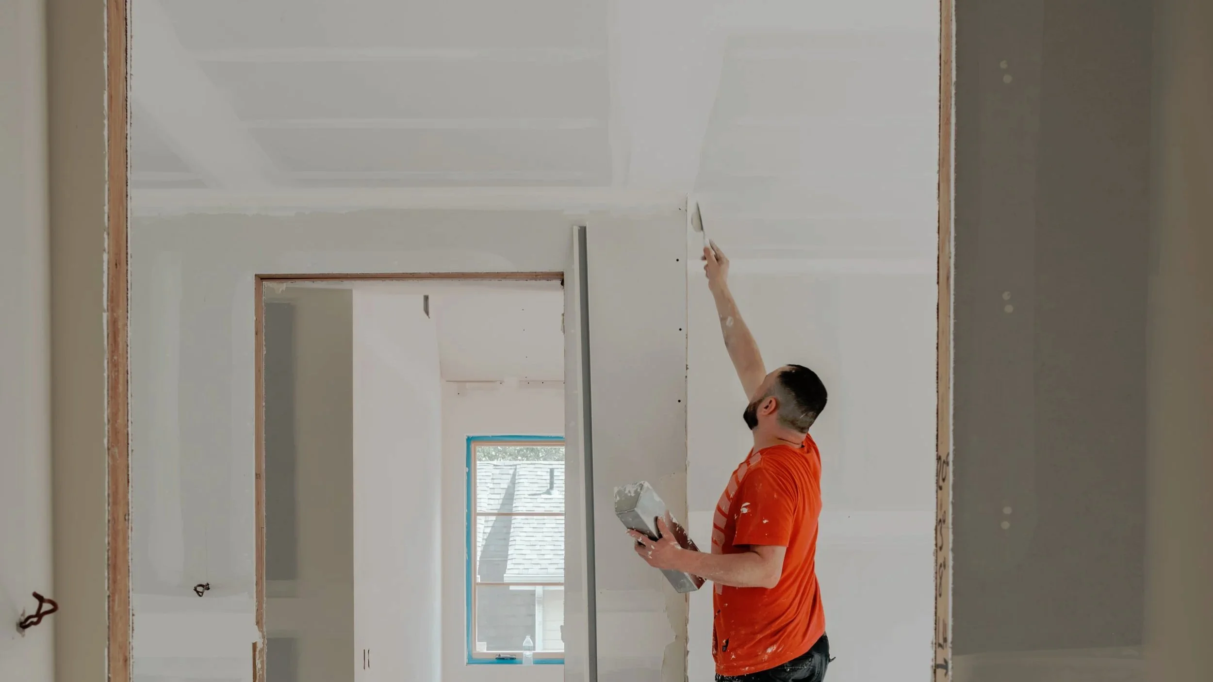A person applying paint to the wall in a room under construction, holding a paintbrush in one hand and a paint container in the other.