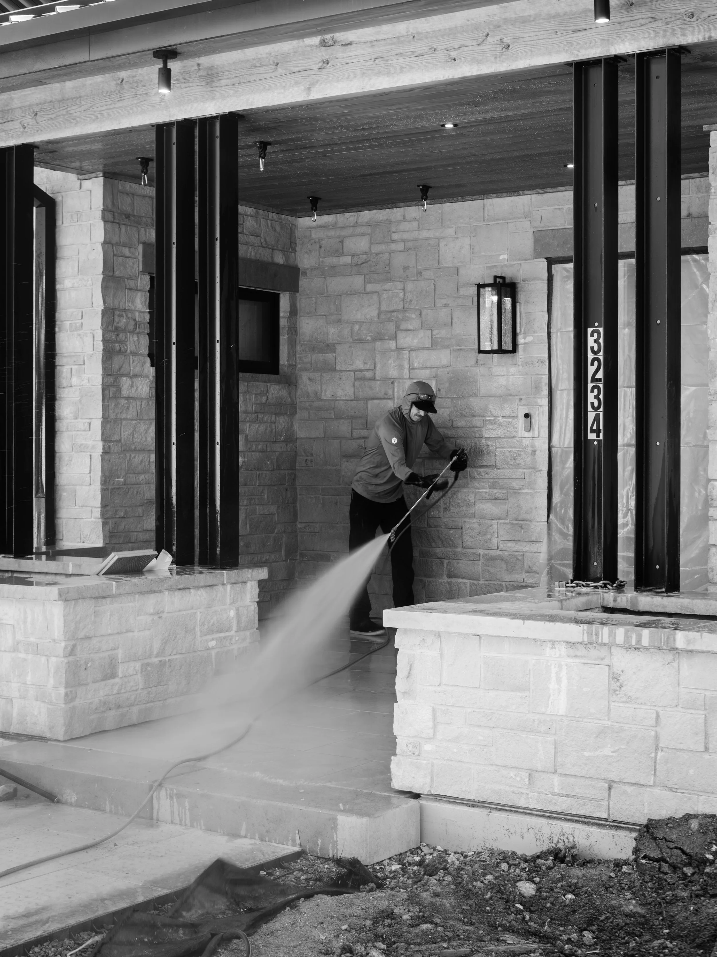 A construction worker wearing a helmet and glasses cleaning the floor at an outdoor building site. The site has a brick wall, black metal columns, and a wooden ceiling with installed lighting fixtures.