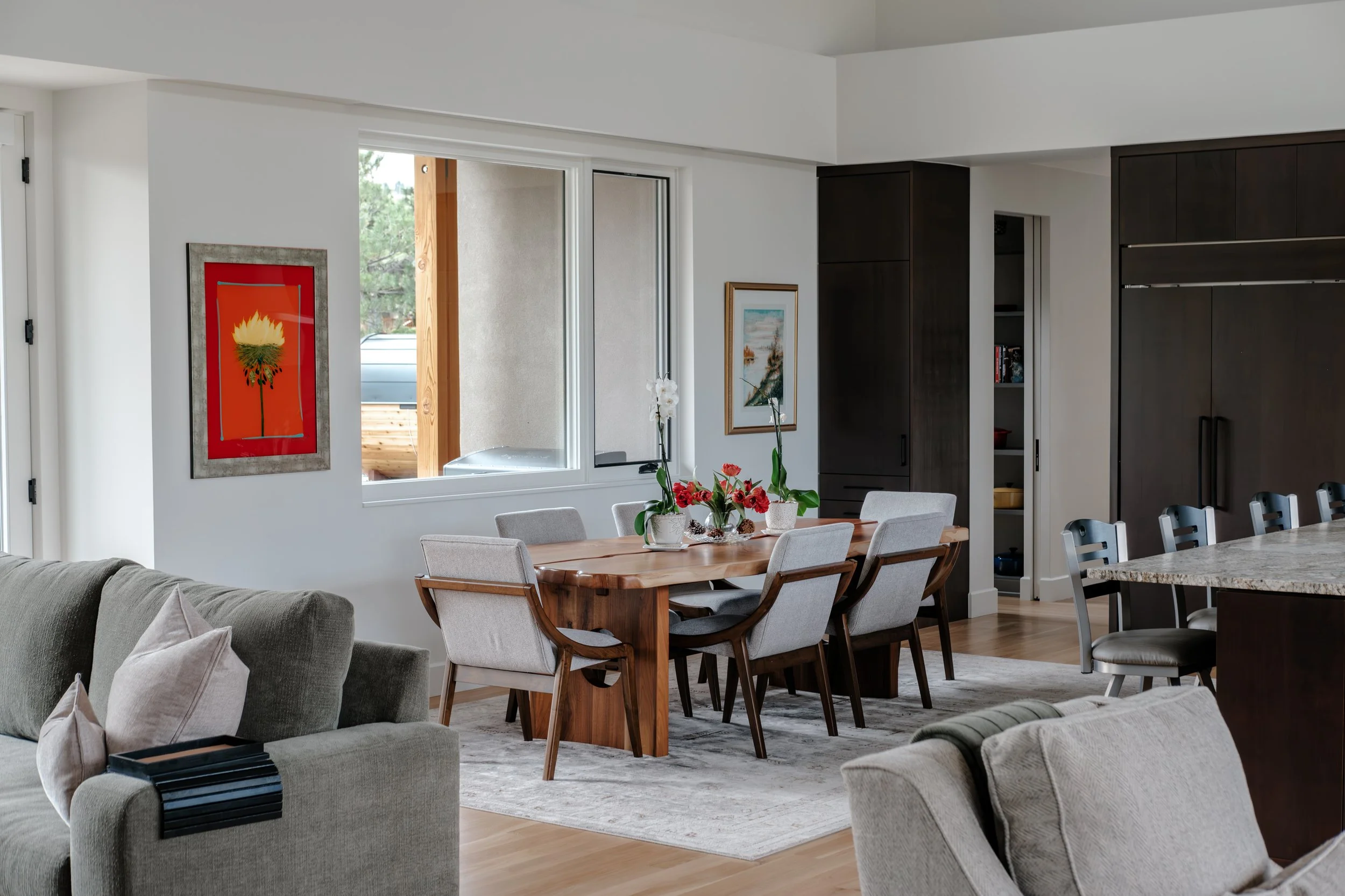 A living and dining area with large windows, a gray sofa with pillows, a wooden dining table with six chairs, and dark wood cabinets, decorated with framed artwork, flowers, and a rug, with a view of outdoor trees.
