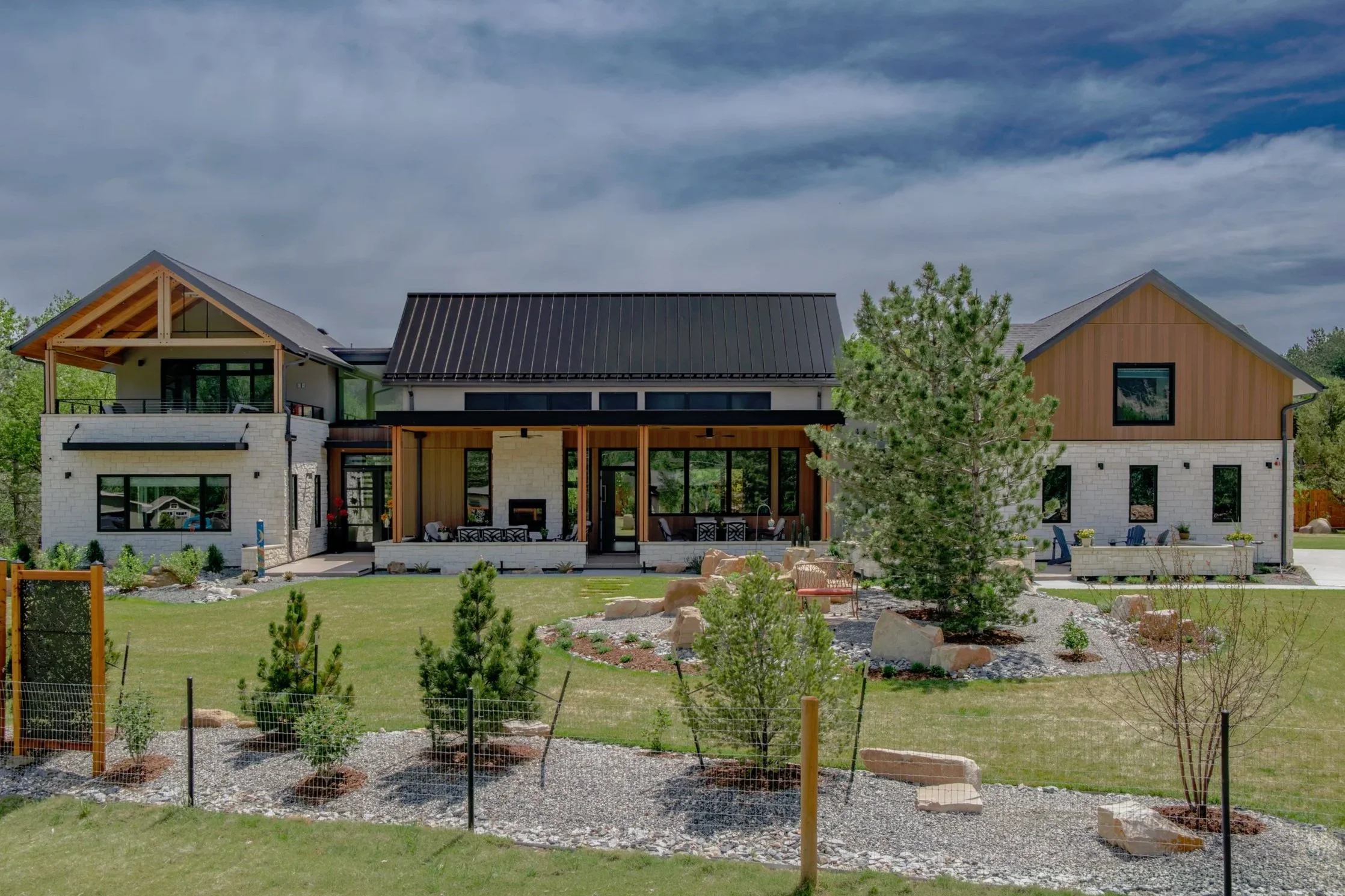 Modern house with a mix of white brick and wooden exterior, black metal roof, large windows, surrounded by a landscaped yard with small trees, rocks, and outdoor seating area under a cloudy sky.