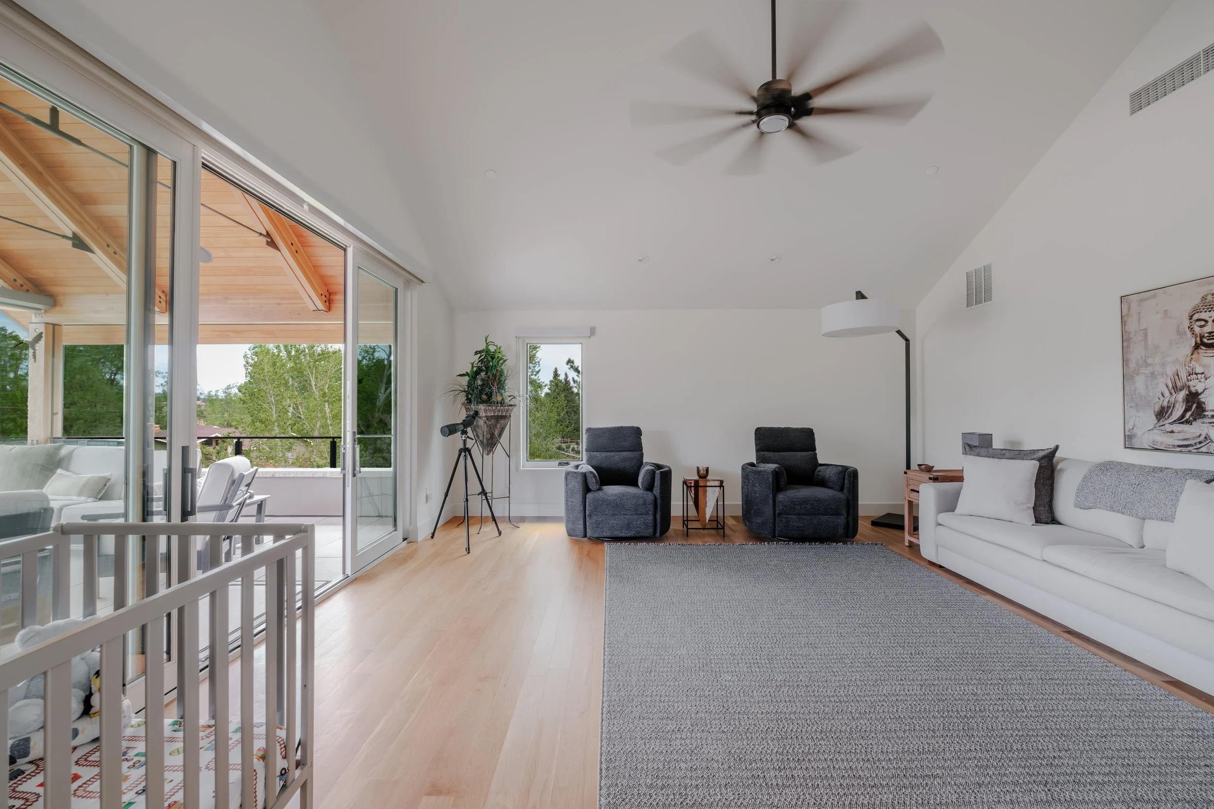 A spacious living room with hardwood floors, two gray armchairs, a white sofa, and a large window with sliding glass doors leading to an outdoor balcony. There is a tripod with a telescope near the window, a painting of a meditative figure on the wal