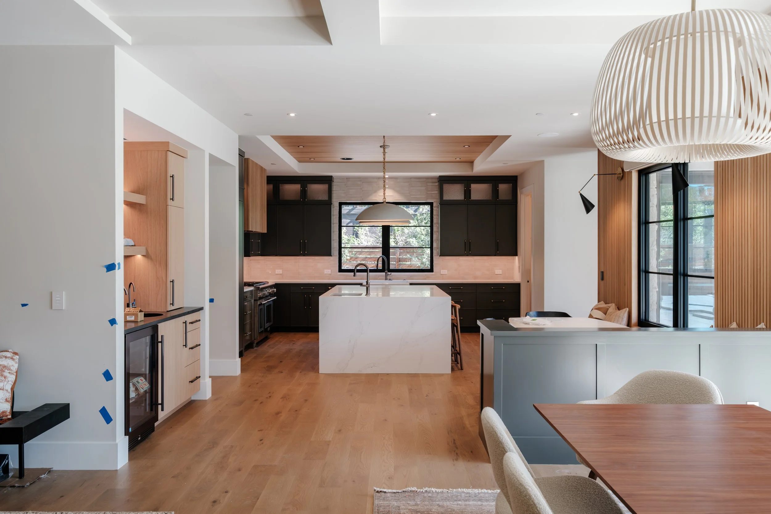 Modern kitchen with wooden cabinets, white island, black cabinetry, large window, and wooden flooring, with a dining table and chairs in the foreground.