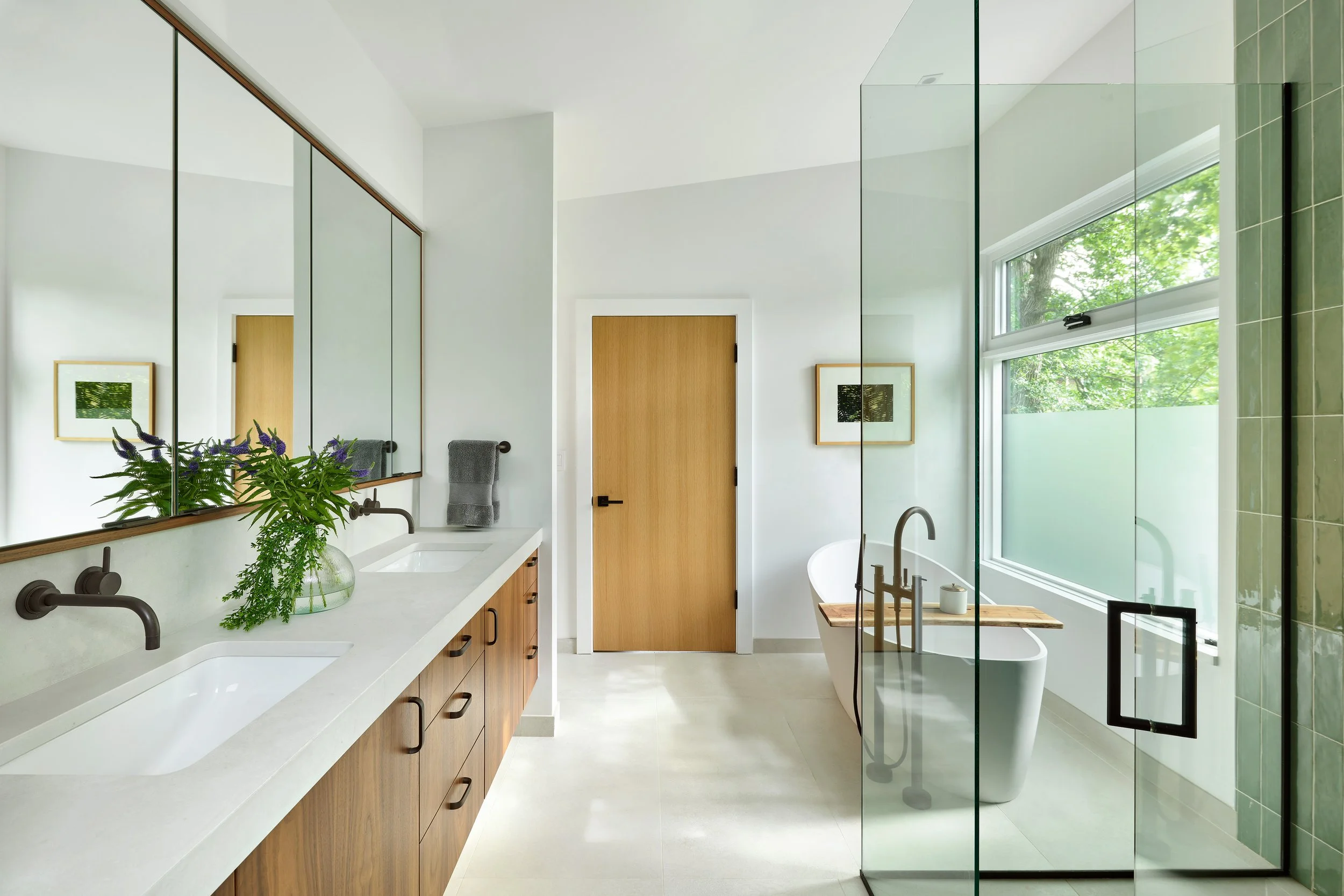 Modern bathroom with double vanity, large mirror, green tile walk-in shower, and window with leafy view