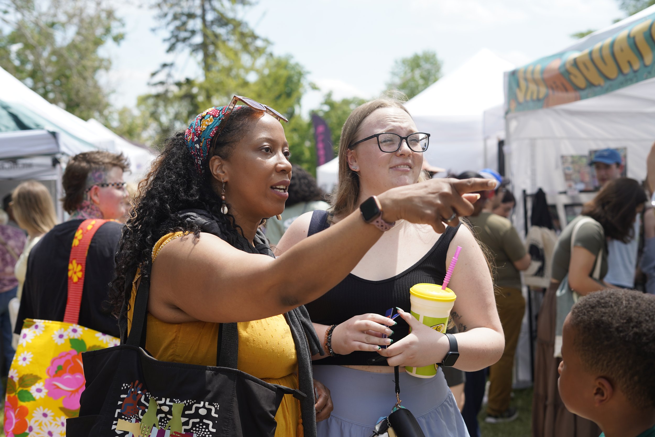Two women at an outdoor event, one wearing a colorful headscarf and sunglasses, pointing with a smile, holding a yellow cup with a pink straw, while the other woman with glasses also smiles and holds a similar drink, surrounded by tents and people.