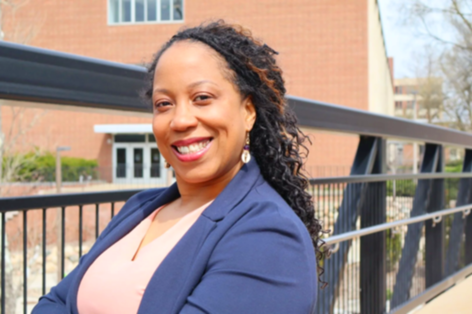 A woman with curly hair wearing a blue blazer and earrings, smiling while sitting outdoors near a black metal railing, with a brick building in the background.