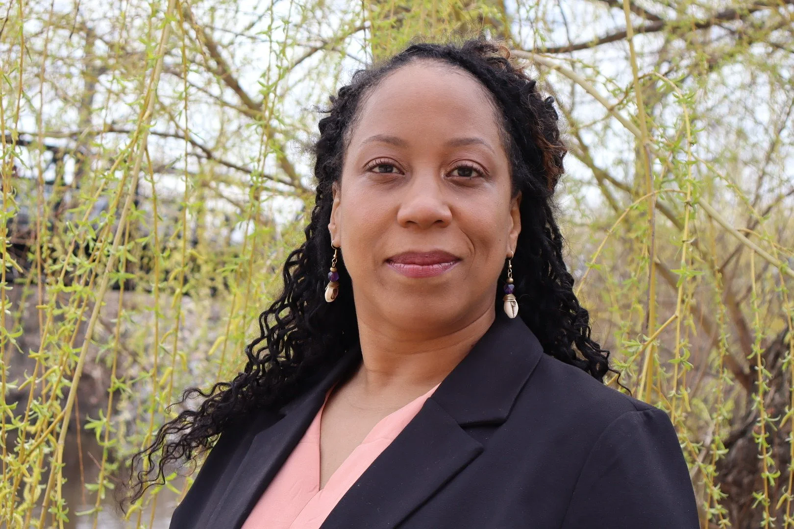 A woman with curly black hair and earrings, wearing a black blazer and a pink blouse, standing outdoors with a background of yellow-green trees.