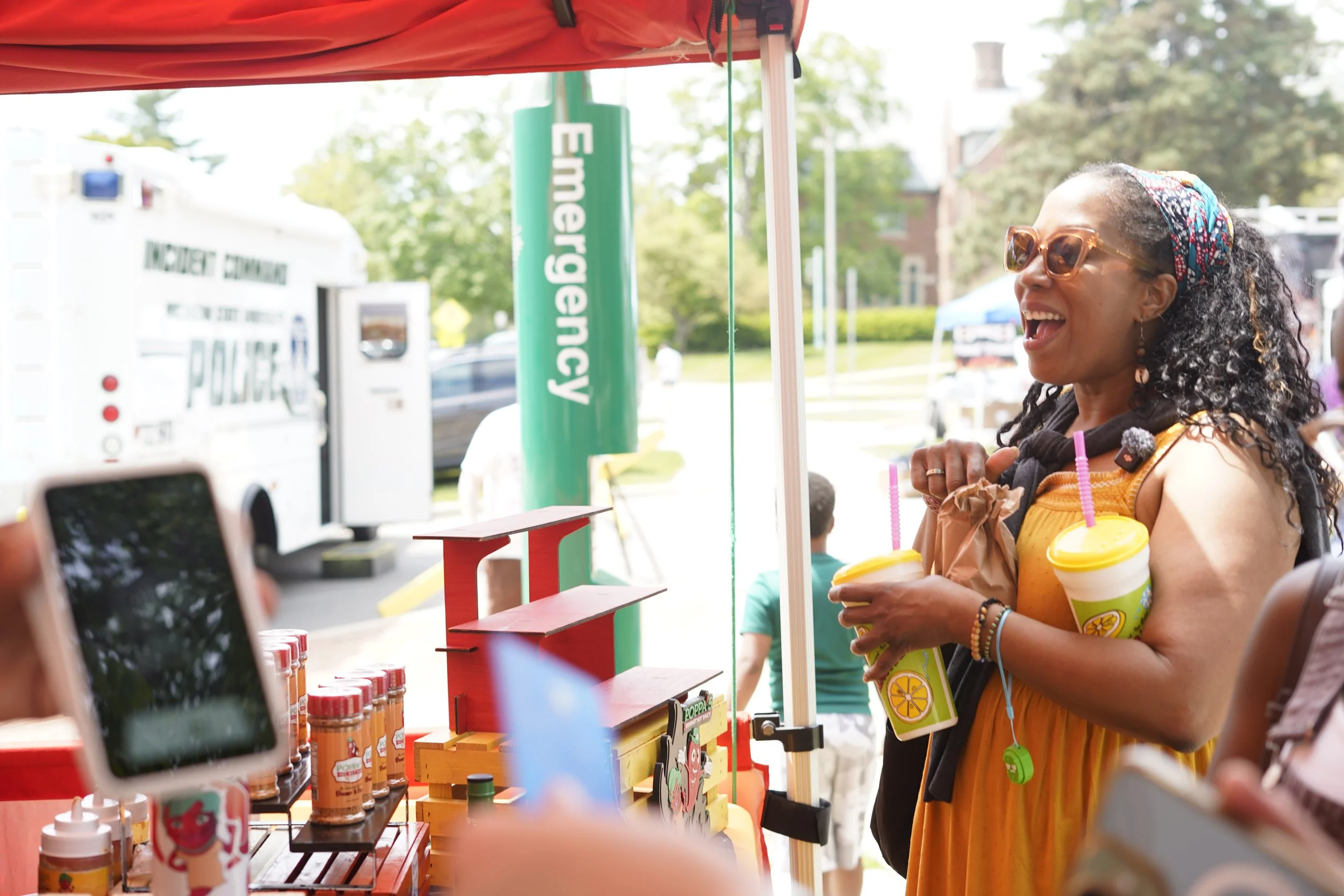A woman laughing and holding two drinks with lemon slices at an outdoor market stall, with a green sign reading 'Emergency' and various bottled products on display.